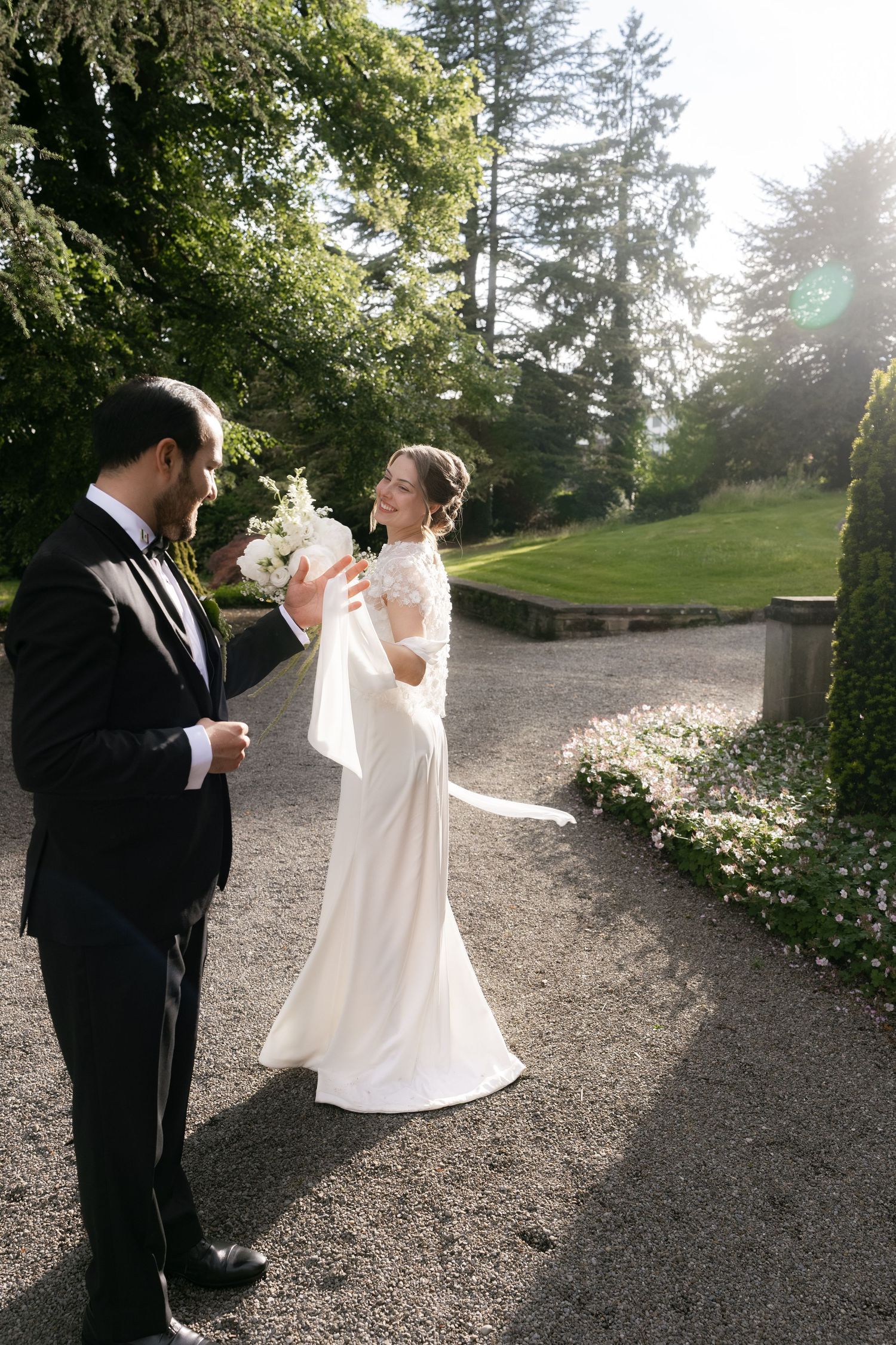 Bride in white gown holds bouquet while groom in black tuxedo turns for first look in lush garden.