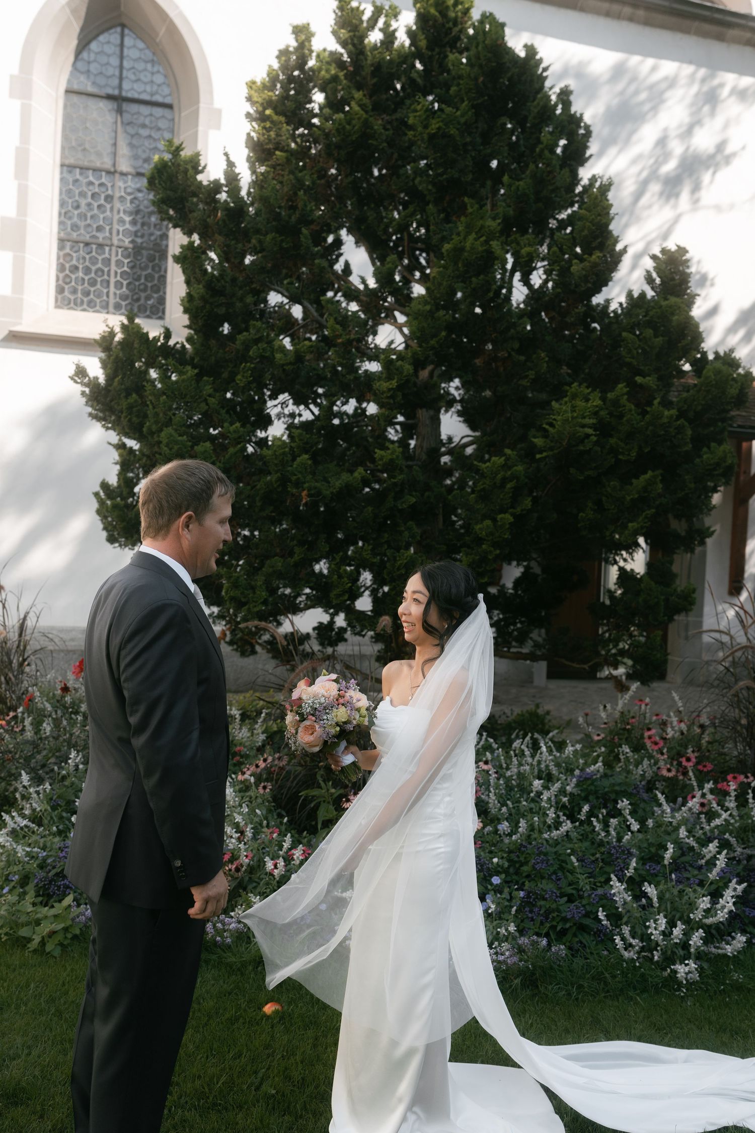 Elegant wedding couple posing in front of a white church with lush garden scenery.