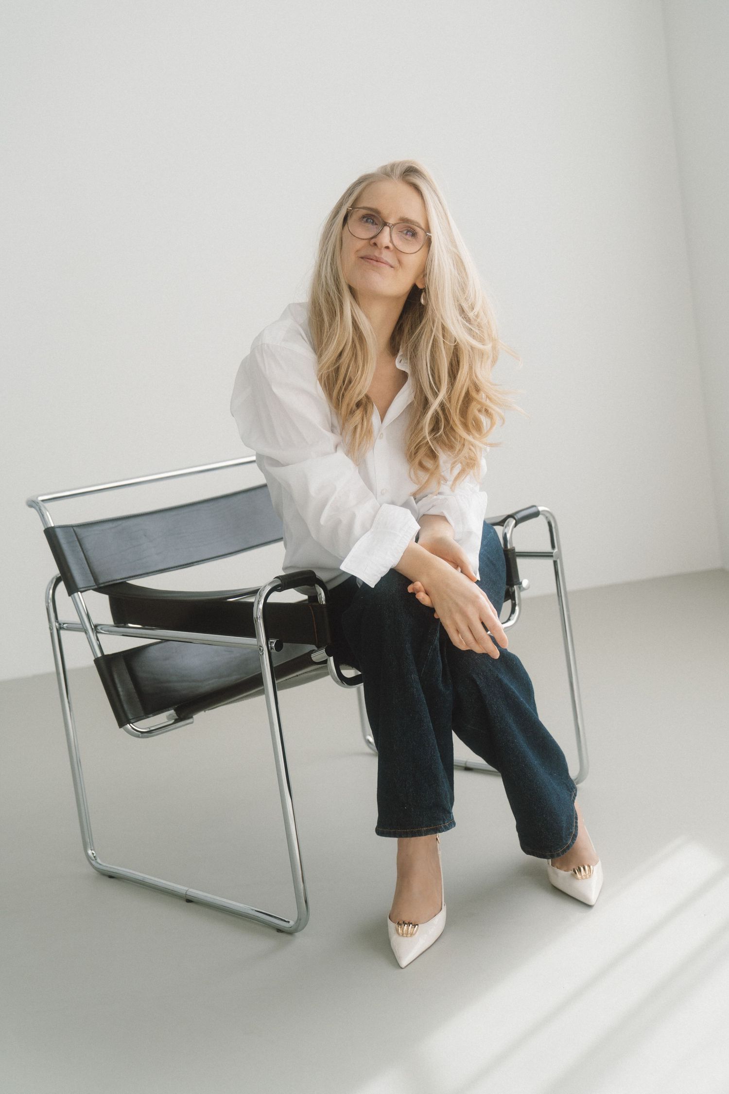 A person in a white shirt and dark pants poses on a modern gray chair in a minimalist white studio setting.