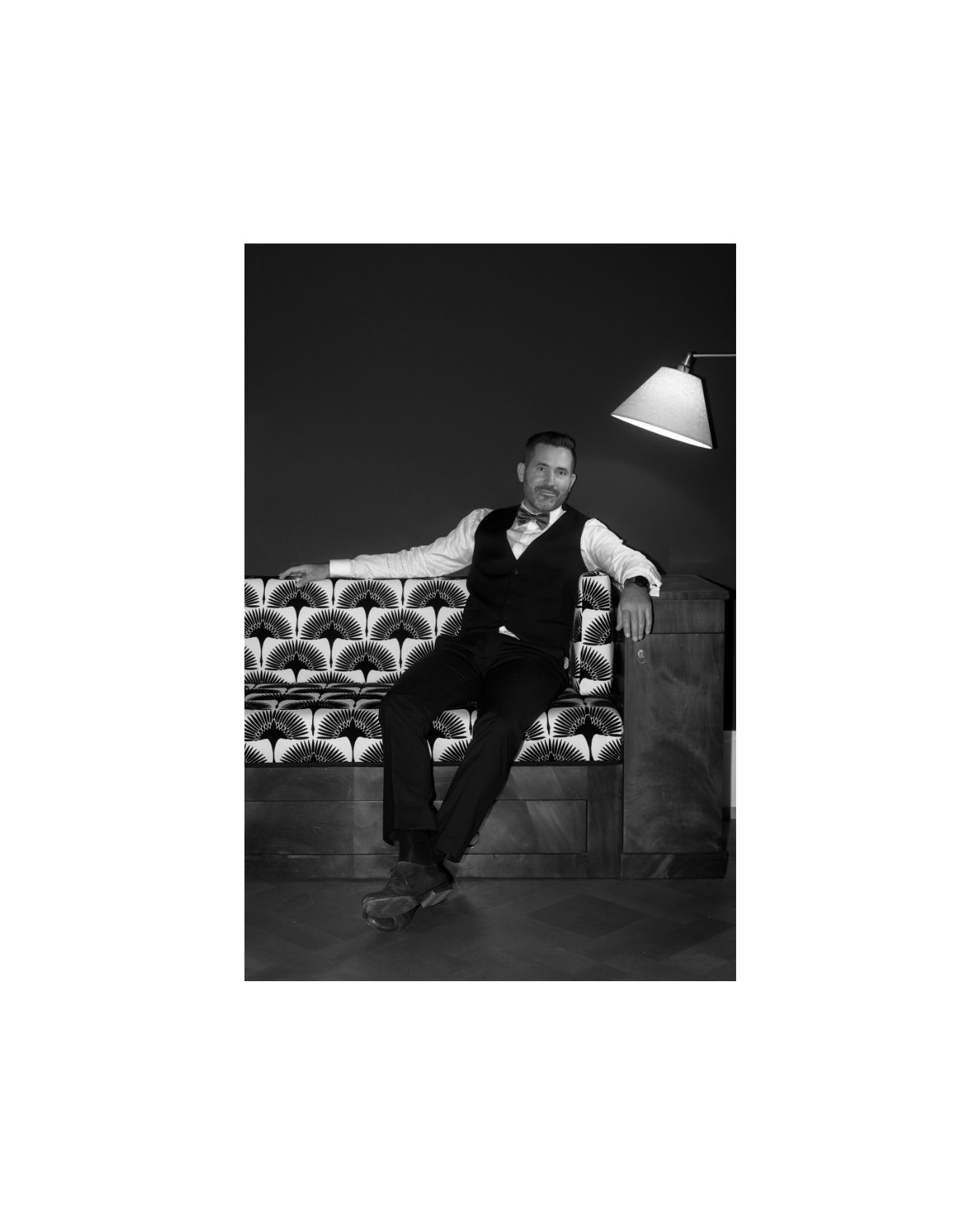 Man in black vest and bow tie seated on patterned sofa beside floor lamp in black and white portrait photo.