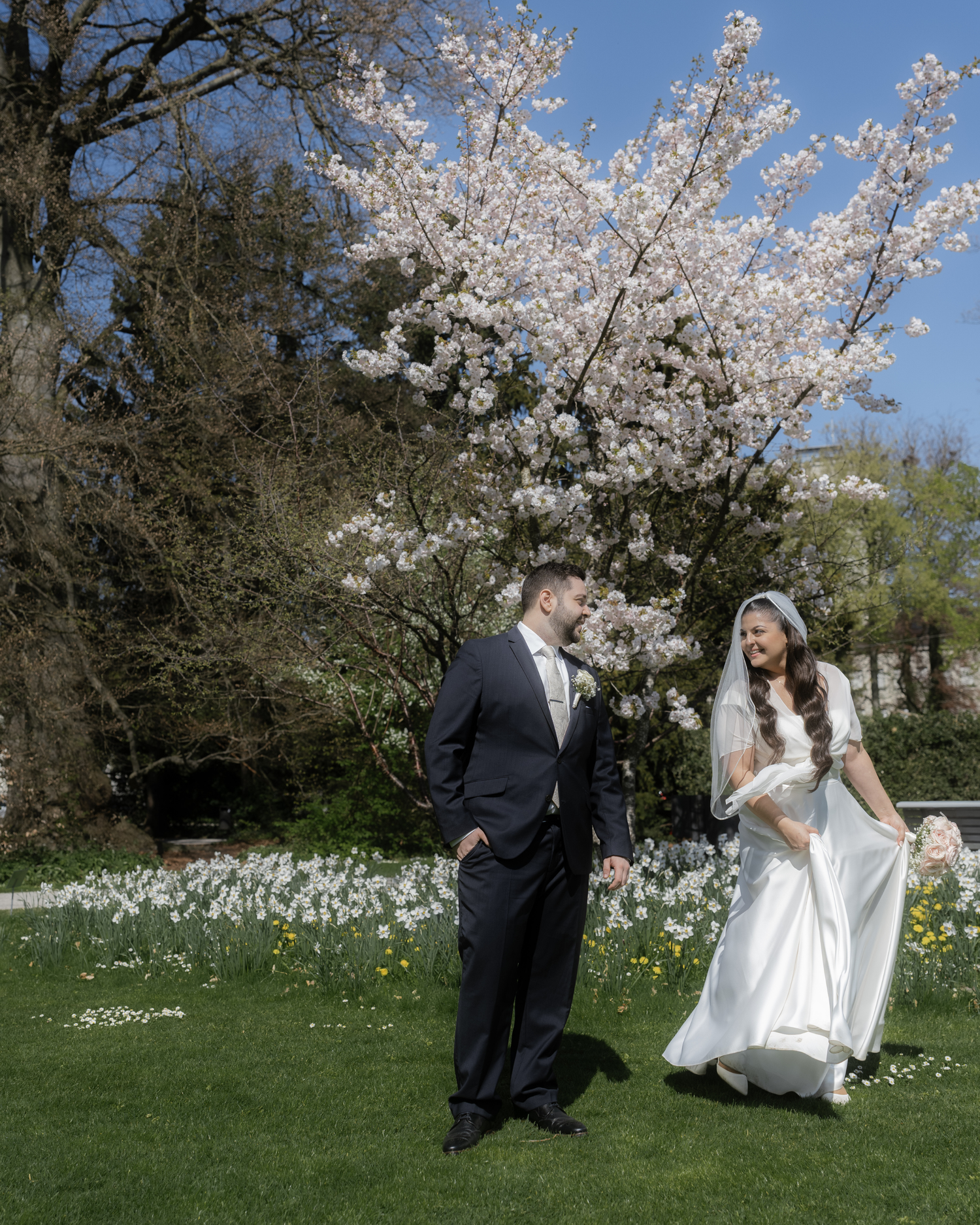 Bride and groom walk hand in hand on lush green lawn beneath blooming cherry blossom tree on sunny spring wedding day.