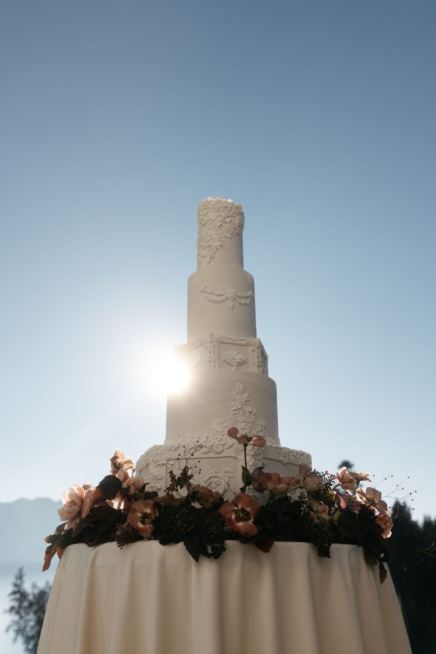 Elegant multi-tiered white wedding cake adorned with floral decorations, backlit by sunlight against a clear blue sky.
