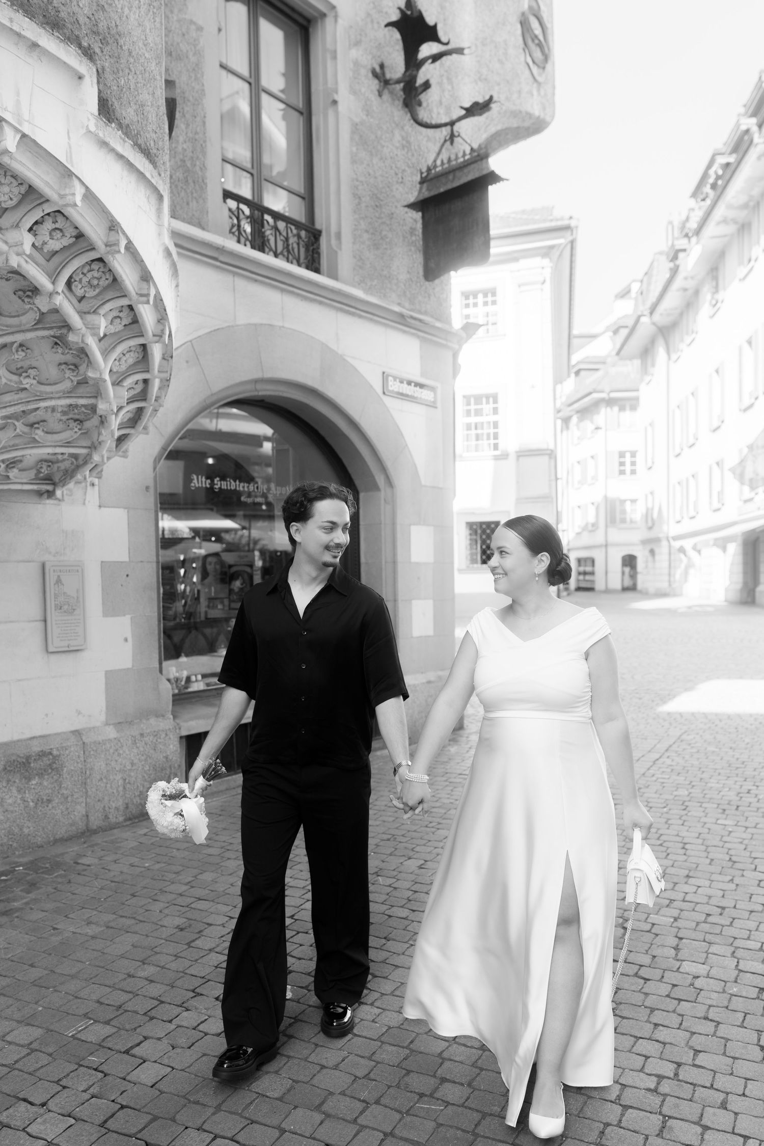 A couple in black and white wedding attire walks together on a cobblestone street in an old European city.