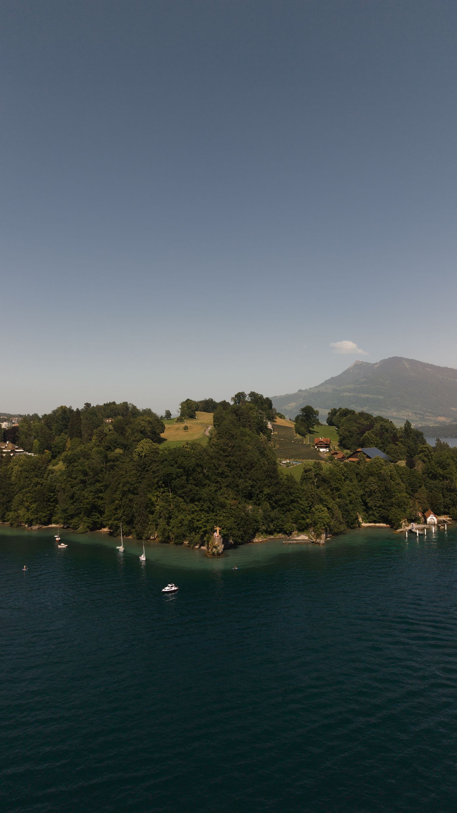 Aerial view of a lush green island peninsula on a serene alpine lake with sailboats and mountains in the background.