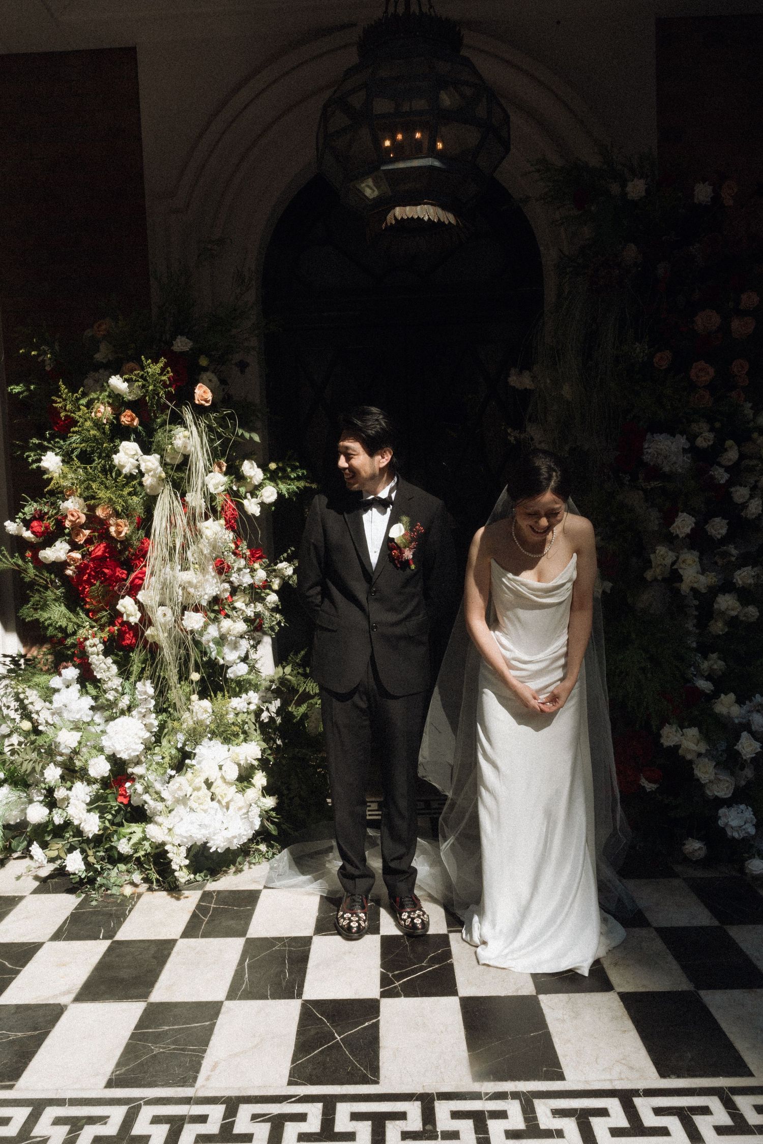 Bride in white gown and groom in black tuxedo stand among lush floral arrangements on a checkered marble floor.