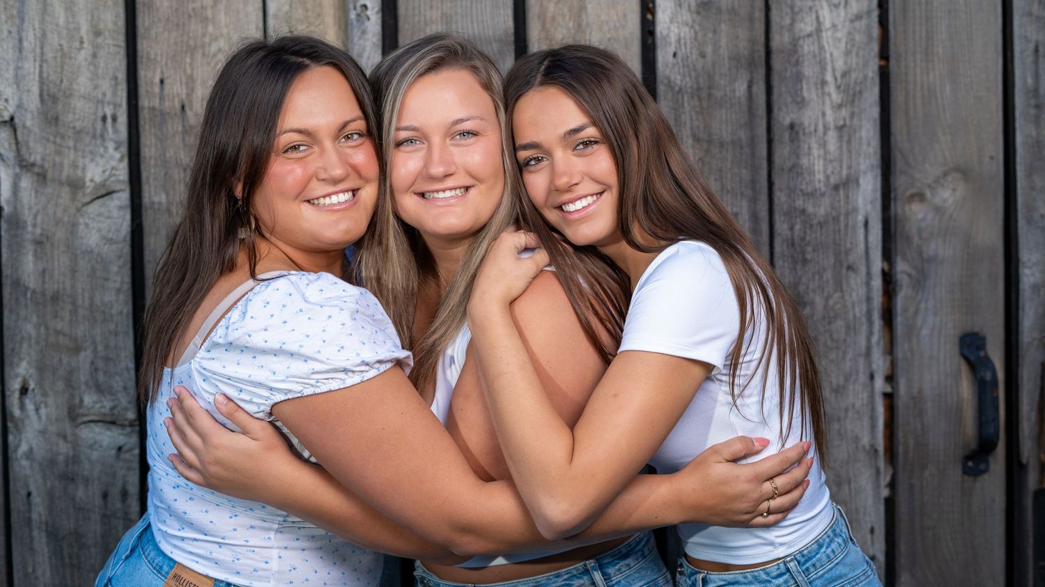 Three sisters in white shirts and jeans embracing outdoors during a portrait session.