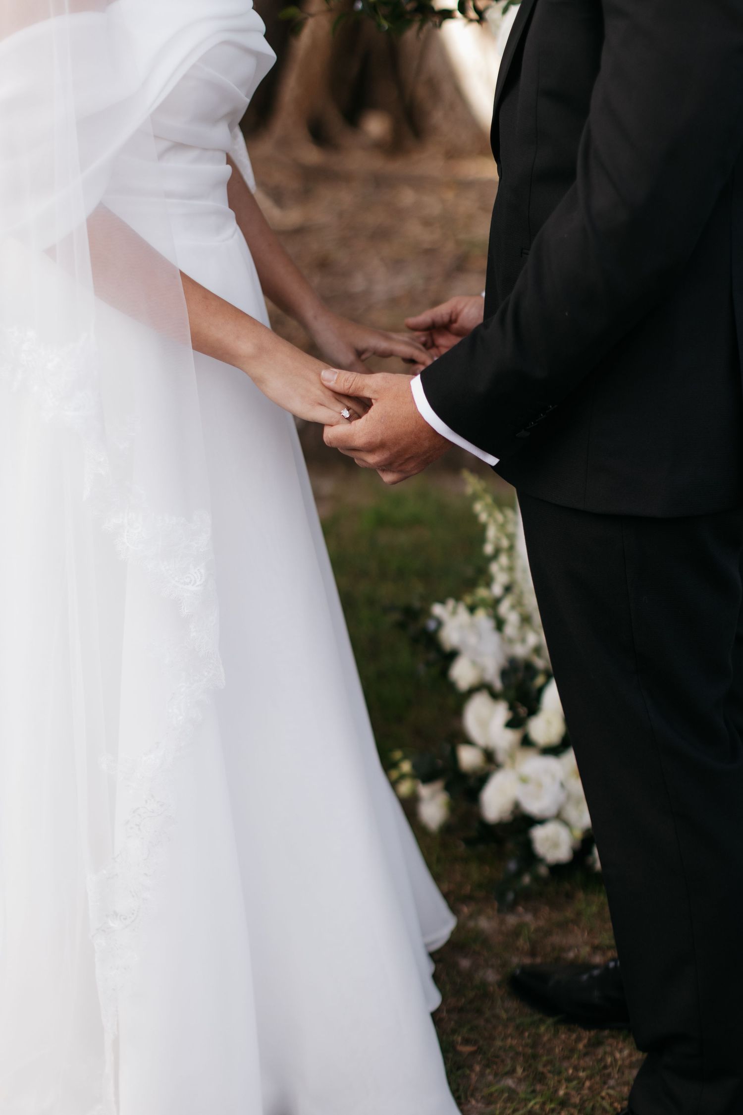 Close-up of bride and groom holding hands during outdoor wedding ceremony with white floral decorations.