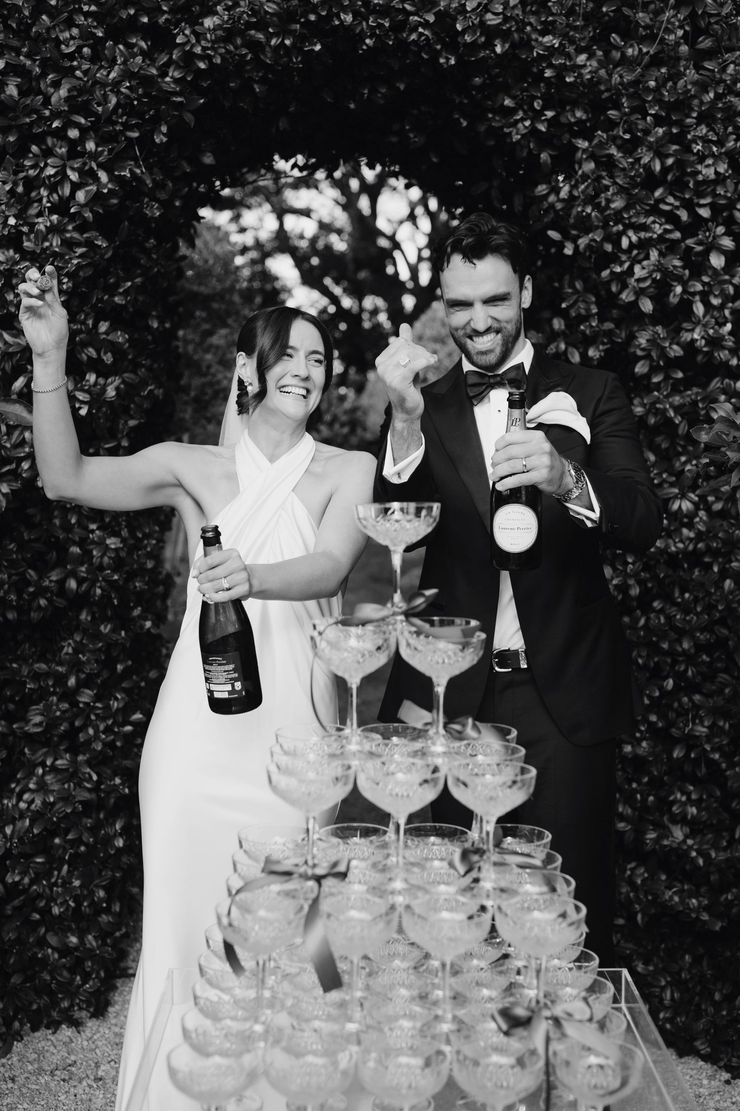 A couple celebrates with champagne at their wedding reception by a champagne tower of coupe glasses in a garden setting.
