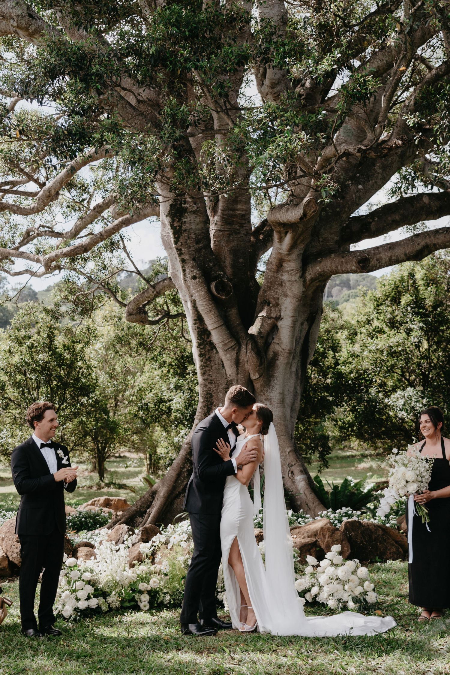 A romantic outdoor wedding ceremony takes place under a majestic old tree adorned with white floral arrangements.