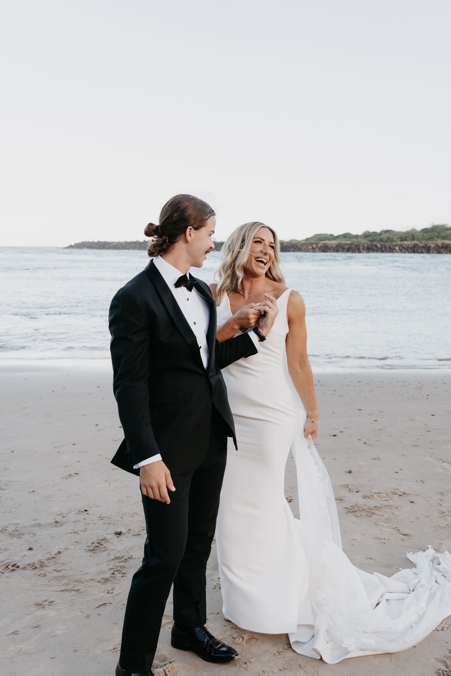 A couple in formal wedding attire shares a joyful moment together on a scenic beach with ocean waves in the background.