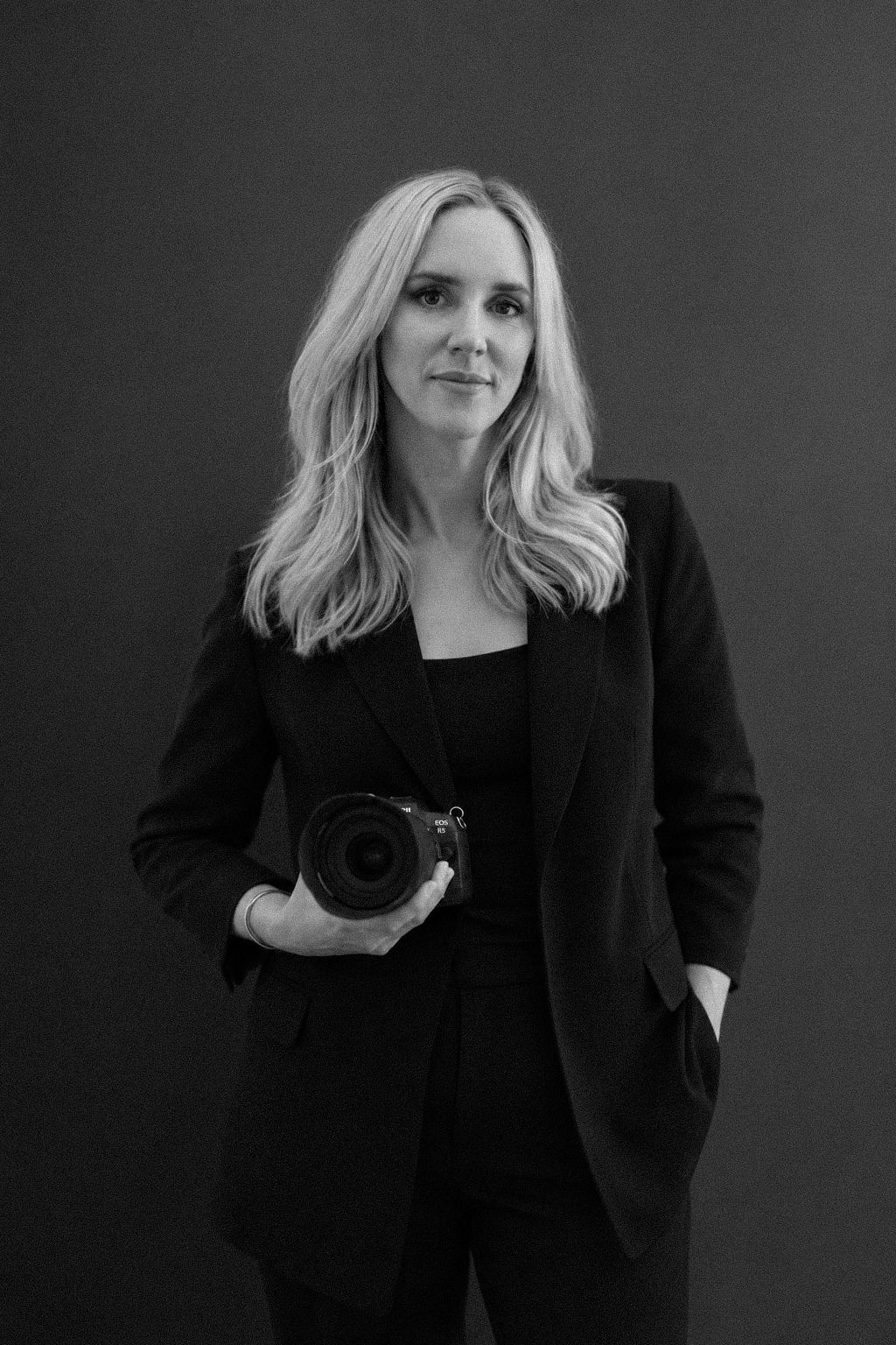 Black and white portrait of a blonde woman in a black blazer holding a professional camera against a dark background.