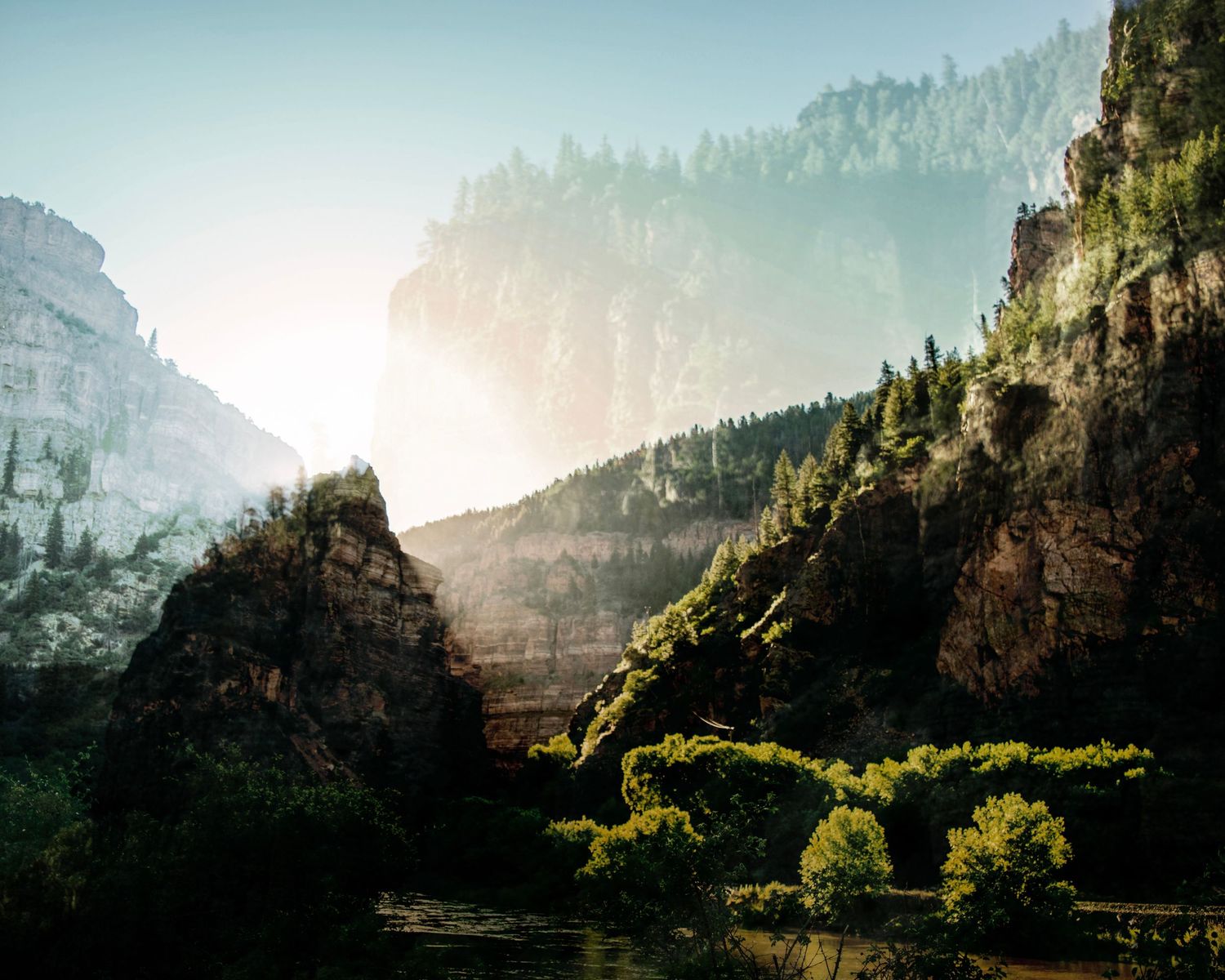 Sunlit canyon with towering rocky cliffs, dense pine trees, and a calm river winding through the misty valley below.