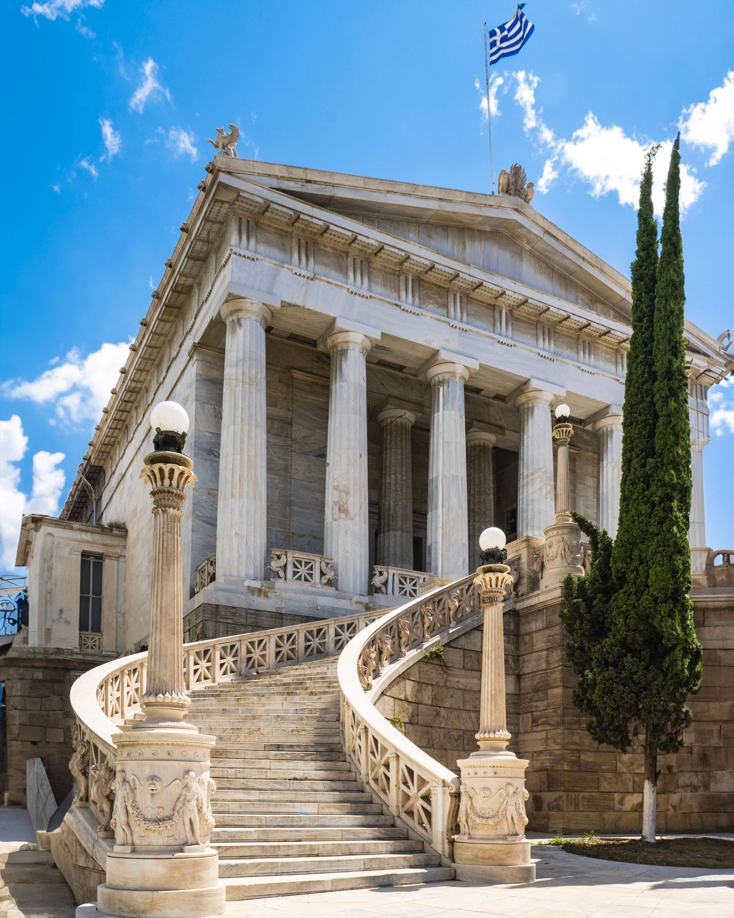 Neoclassical Greek building with marble columns, ornate curved staircase, and Greek flag under blue sky.