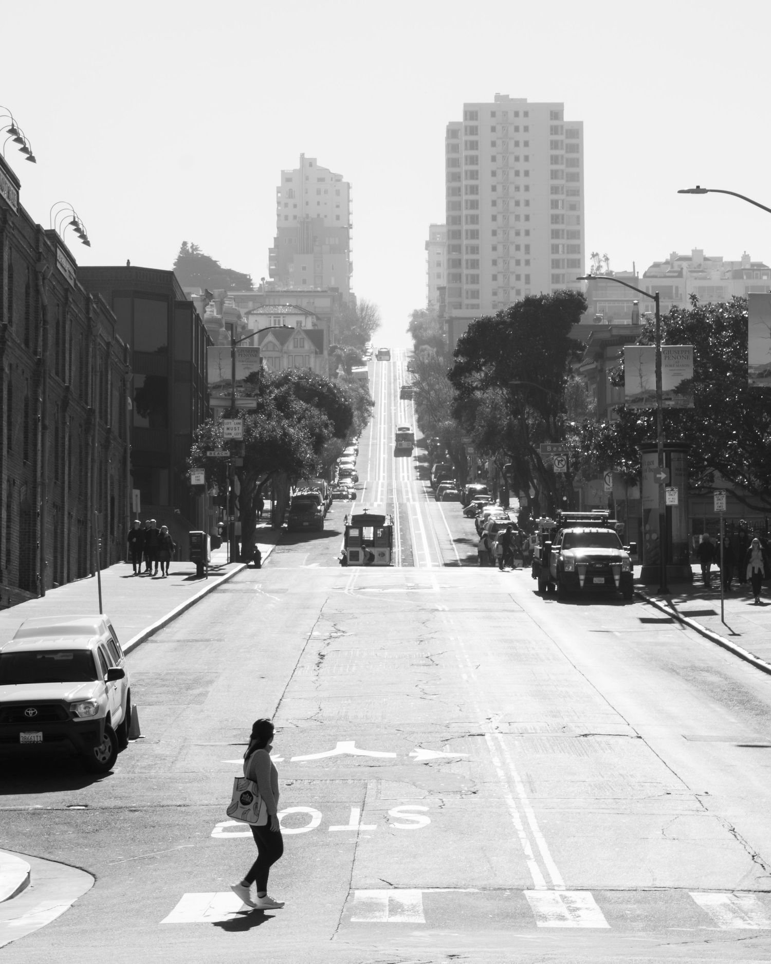 Black and white photo of a woman crossing a steep San Francisco street with city buildings in the background.