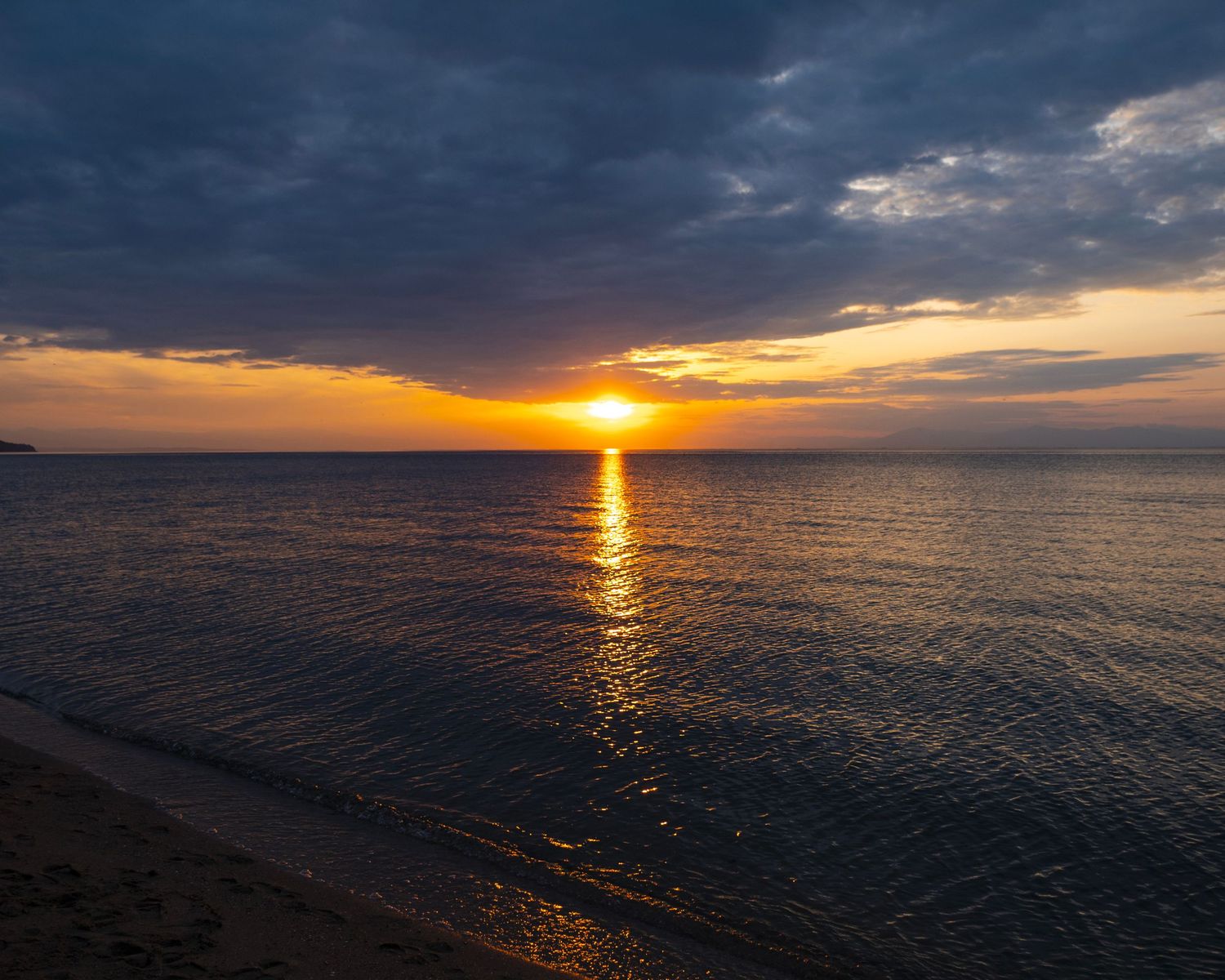 Golden sunset over calm ocean waters with dramatic cloudy sky and sandy beach shoreline in the foreground.