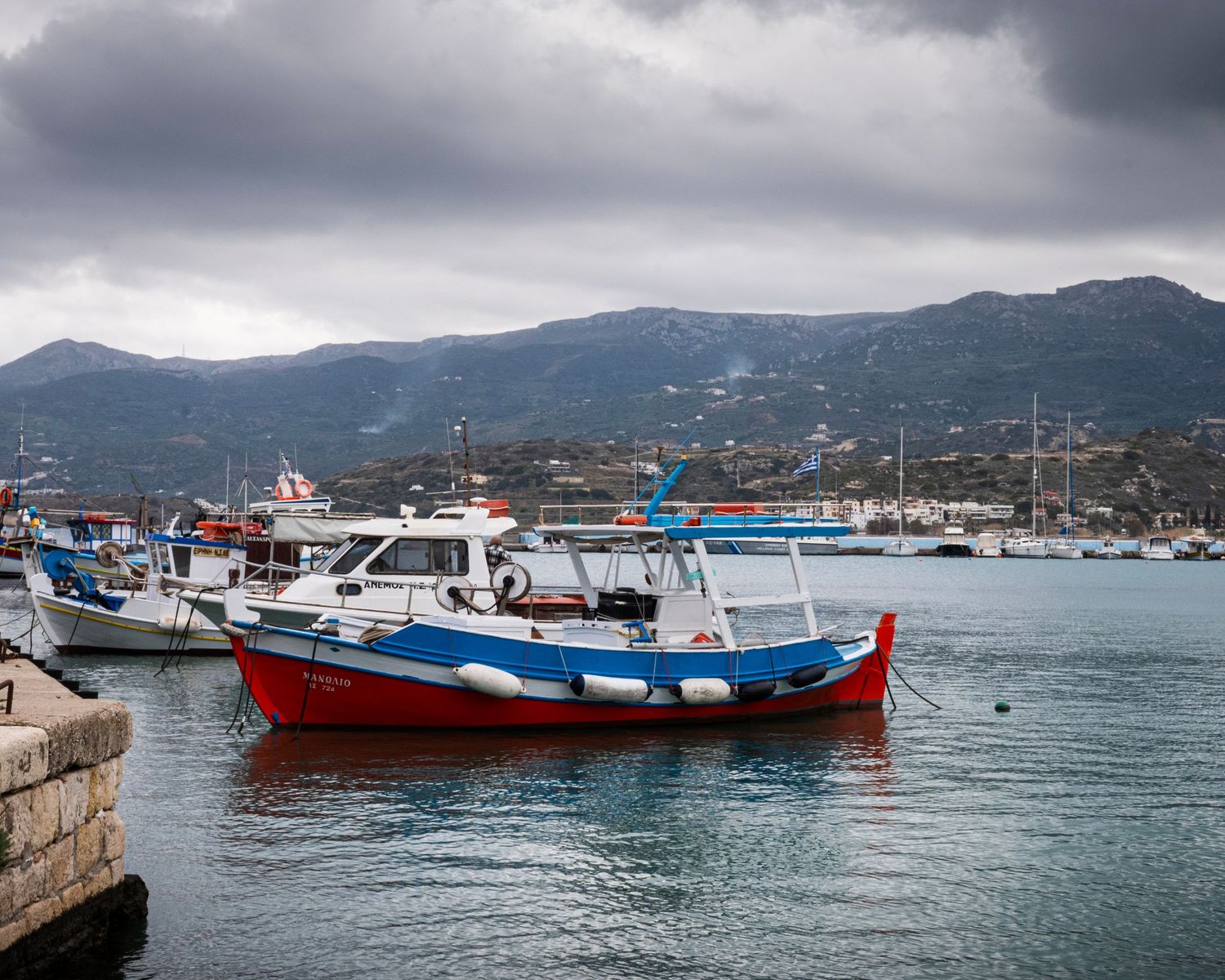 Colorful fishing boats docked at a Mediterranean harbor with mountains and cloudy skies in the background.
