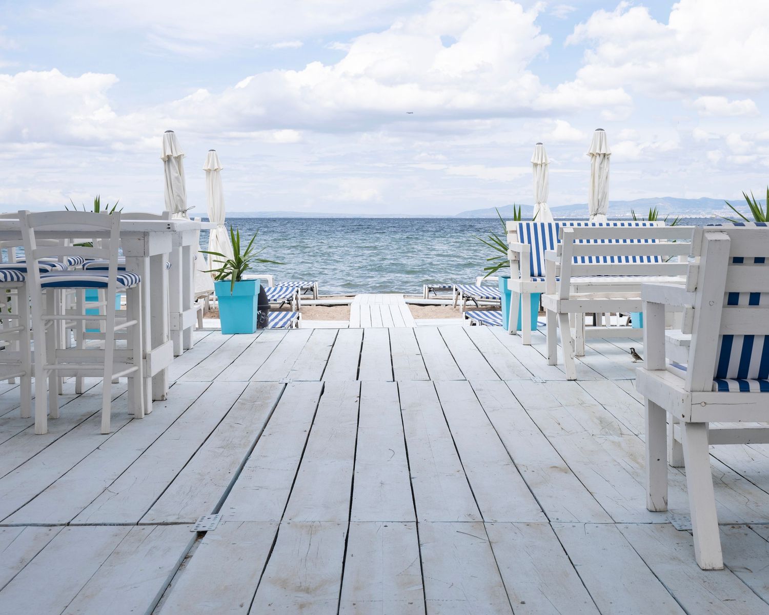 White wooden deck overlooking calm blue ocean with white chairs and blue striped umbrellas on a cloudy day.
