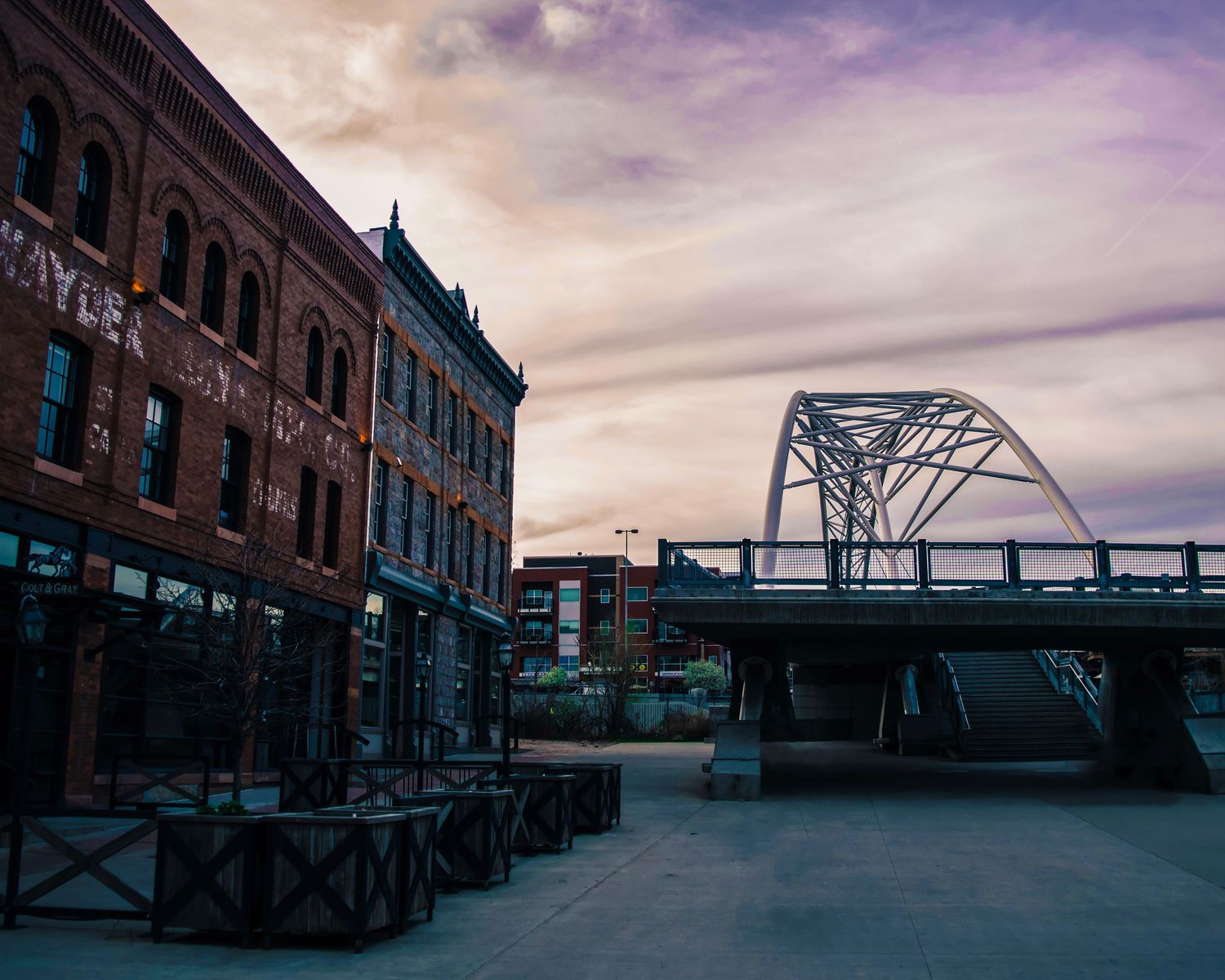 Historic brick buildings line a street beside a steel arch bridge at dusk with dramatic purple clouds overhead.