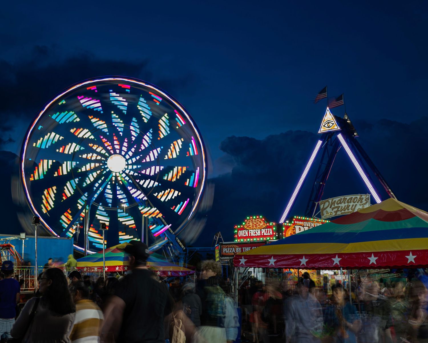 Colorful ferris wheel and carnival rides light up the night sky at a busy outdoor fair with crowds of people.
