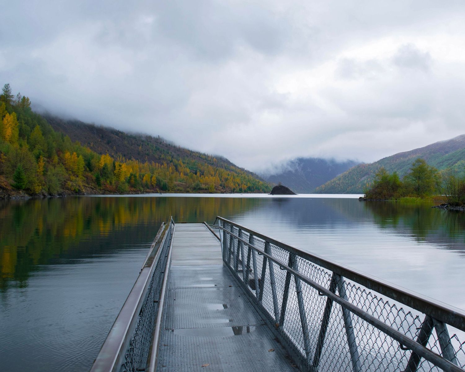 A wet dock extends over a calm mountain lake surrounded by autumn foliage under a cloudy sky.