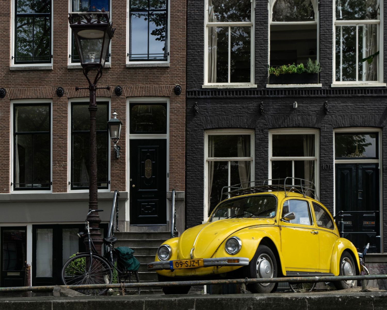 A bright yellow vintage Volkswagen Beetle parked on a canal street in Amsterdam beside classic brick buildings.