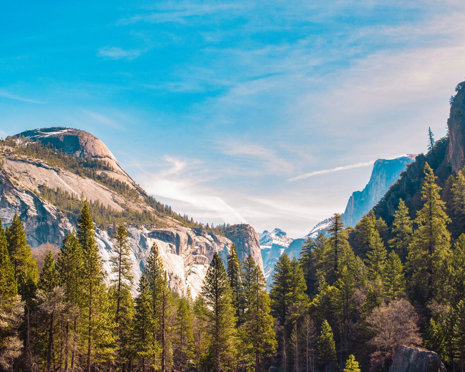Panoramic view of Yosemite Valley with granite peaks, snow-dusted cliffs, and dense pine forests under a blue sky.