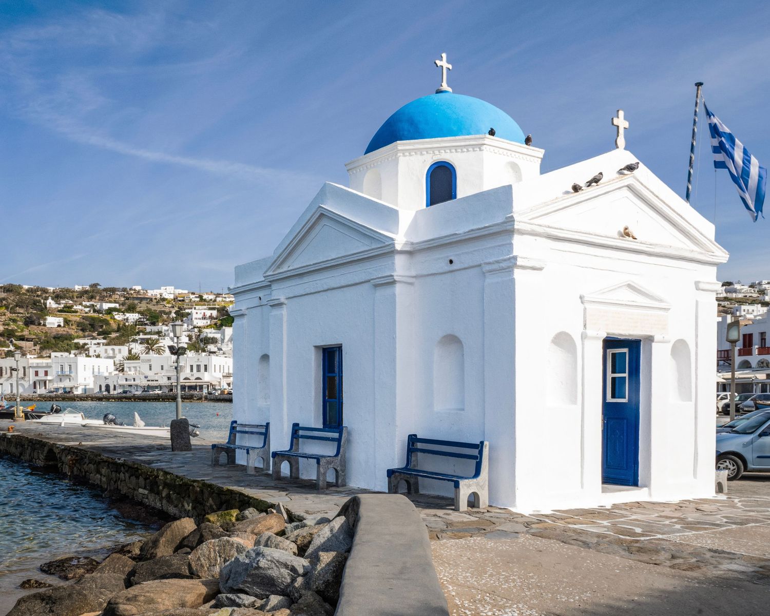 White Greek Orthodox chapel with blue dome and Greek flag by the sea in Mykonos, Greece.