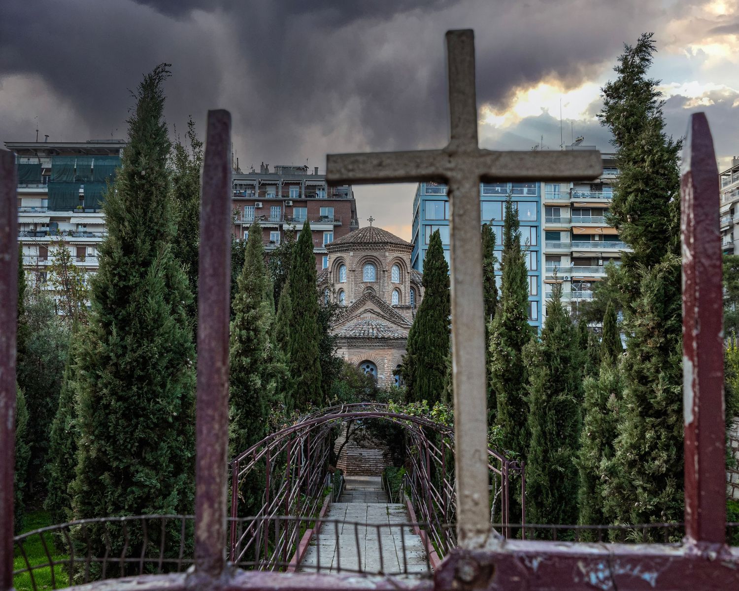 A wooden cross frames a Byzantine church amid cypress trees and city buildings under dramatic stormy skies.