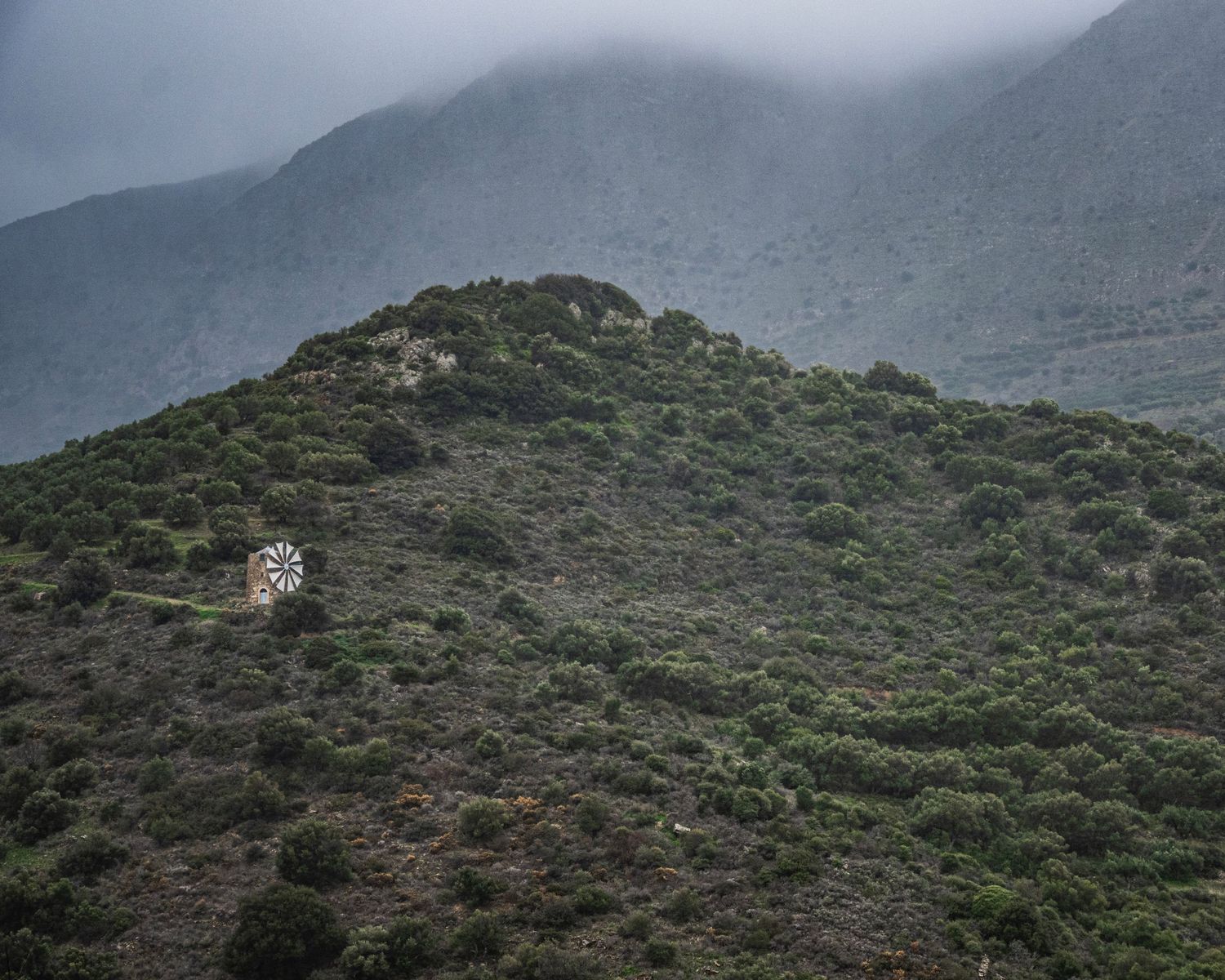 A lone windmill stands on a lush green hillside with misty mountains in the background.