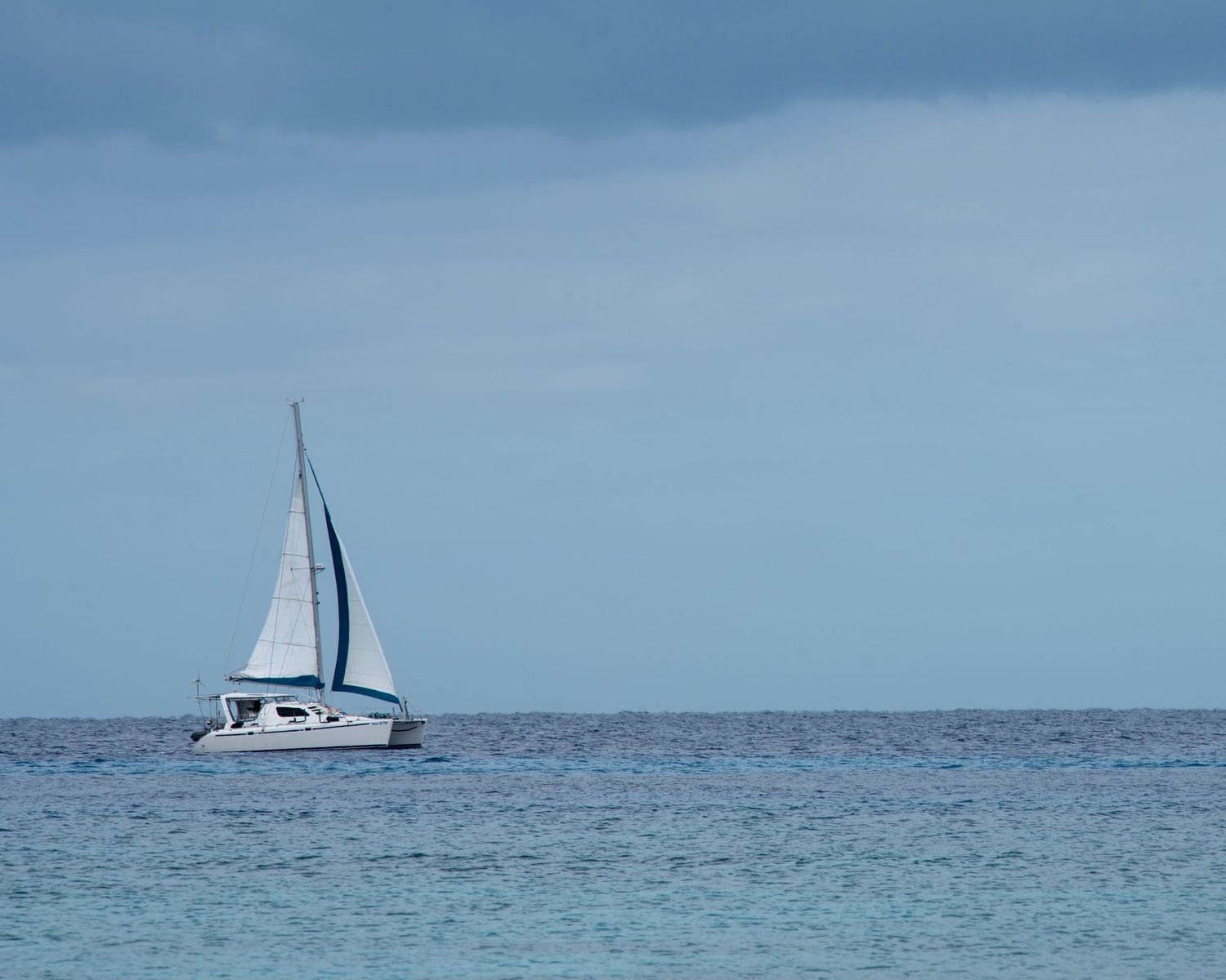 A white sailboat glides on calm blue ocean waters under a partly cloudy sky.