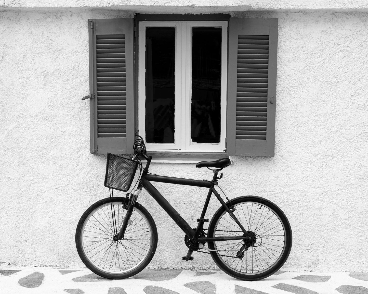 Black and white photo of a bicycle with basket parked against a white stucco wall beside a shuttered window.