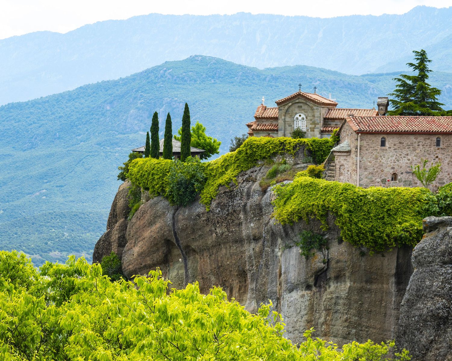 Ancient monastery perched atop rocky cliffs at Meteora, Greece, surrounded by lush greenery and mountain views.