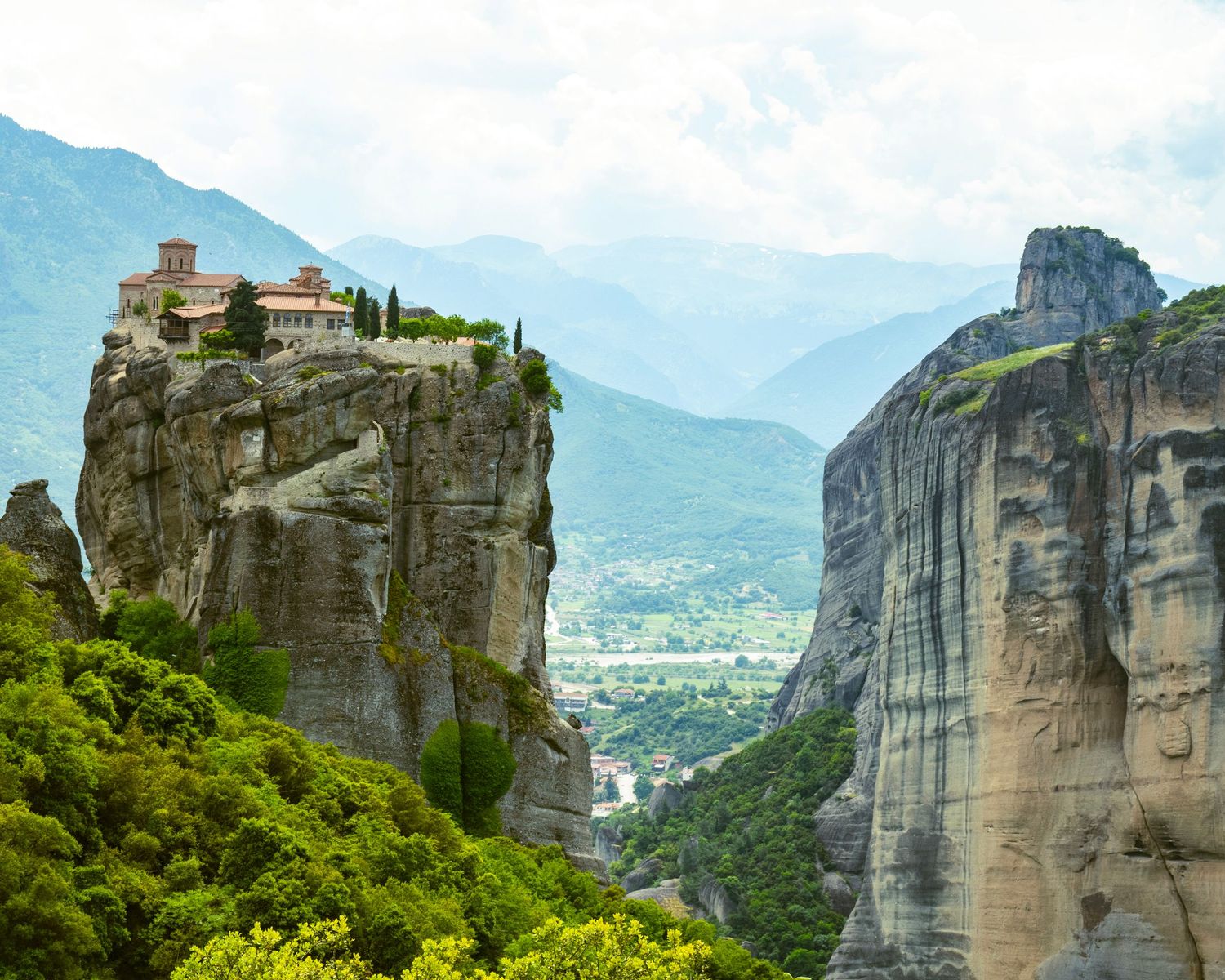 Ancient monastery perched atop dramatic rocky cliffs at Meteora, Greece, surrounded by lush greenery and mountain views.