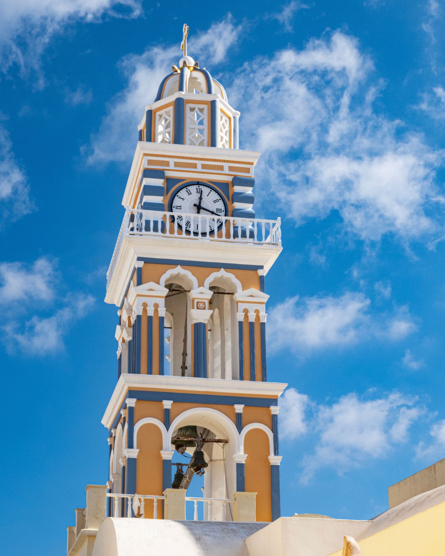 Colorful clock tower with blue and orange stripes, arched bells, and white balconies against a bright blue sky in Santorini, Greece.