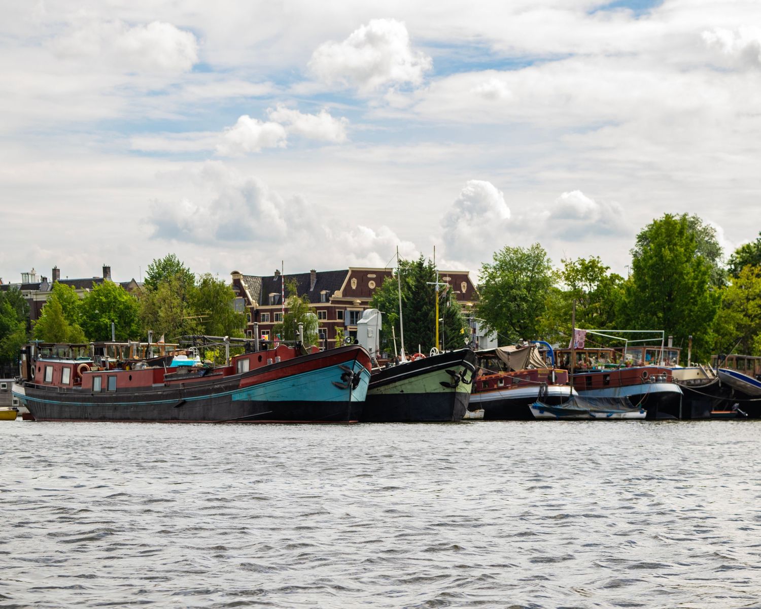 Colorful houseboats moored along an Amsterdam canal with lush green trees and historic buildings in the background.