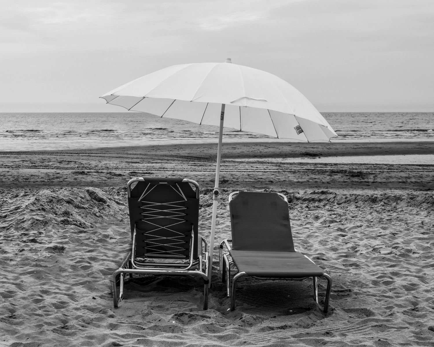 Two empty beach chairs and a white umbrella on a sandy shore with calm ocean waves in the background.