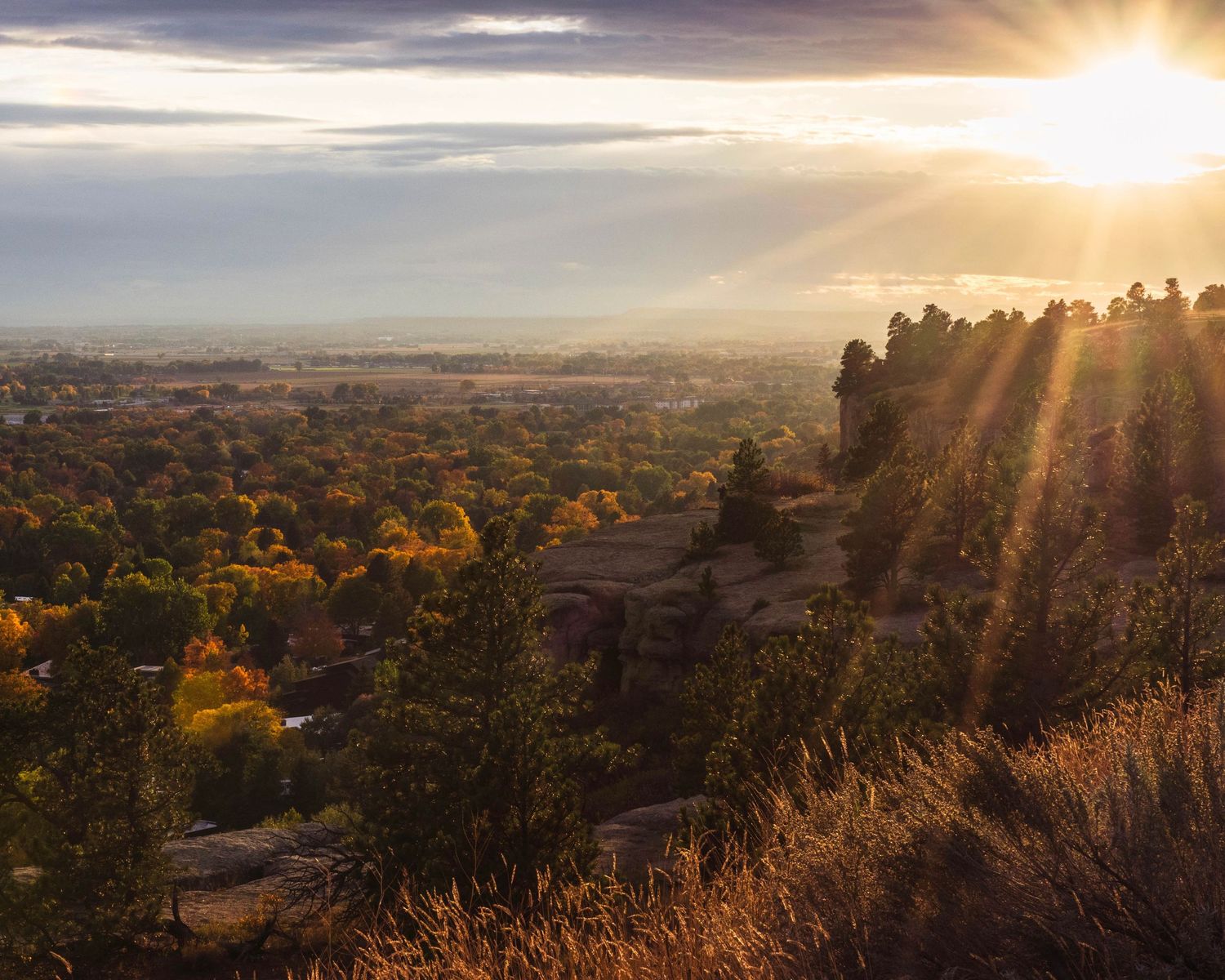 Stunning sunset over a vast autumn landscape with golden trees, rocky terrain, and sun rays bursting through dramatic clouds.