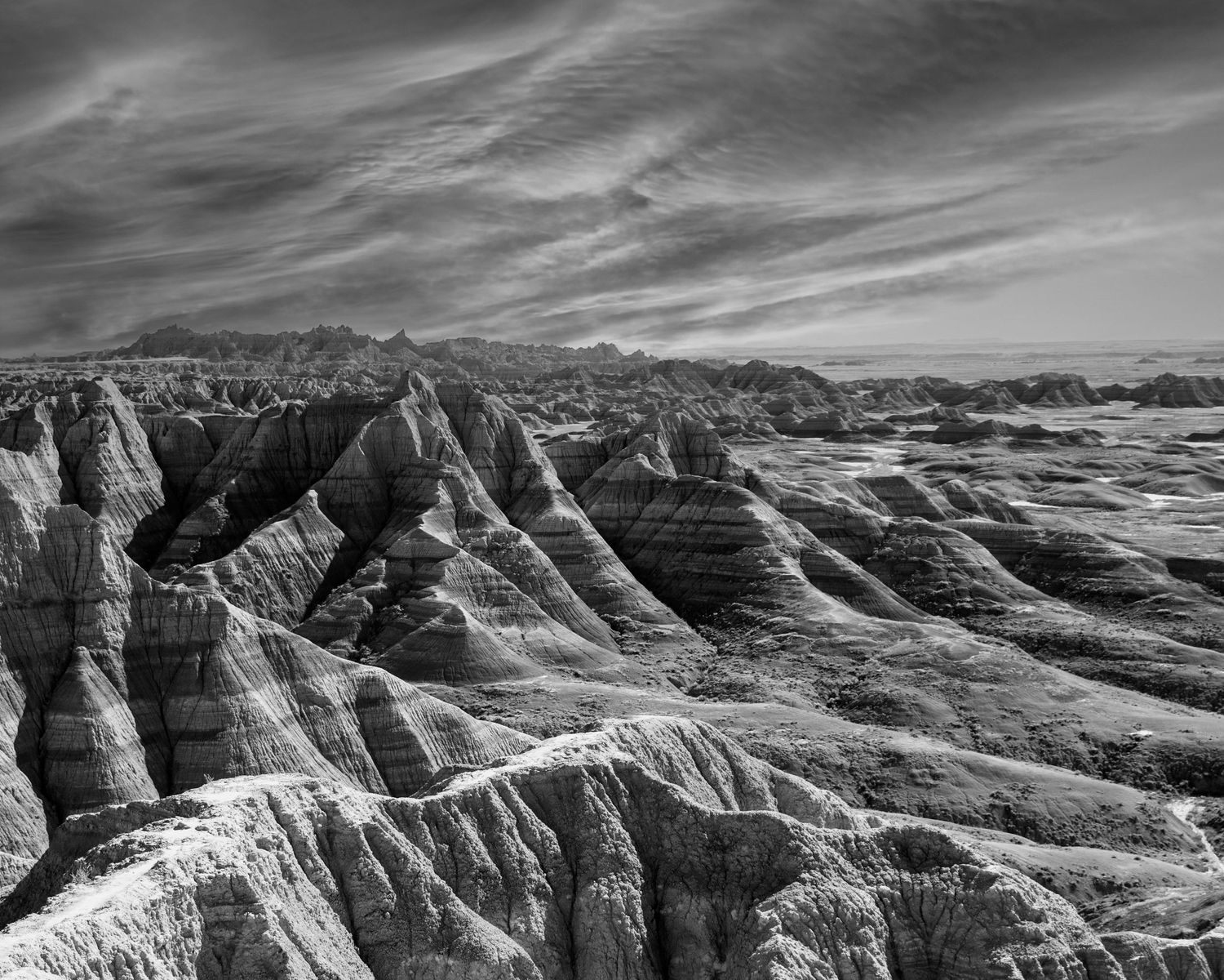Black and white aerial view of eroded badlands rock formations under dramatic cloudy sky.