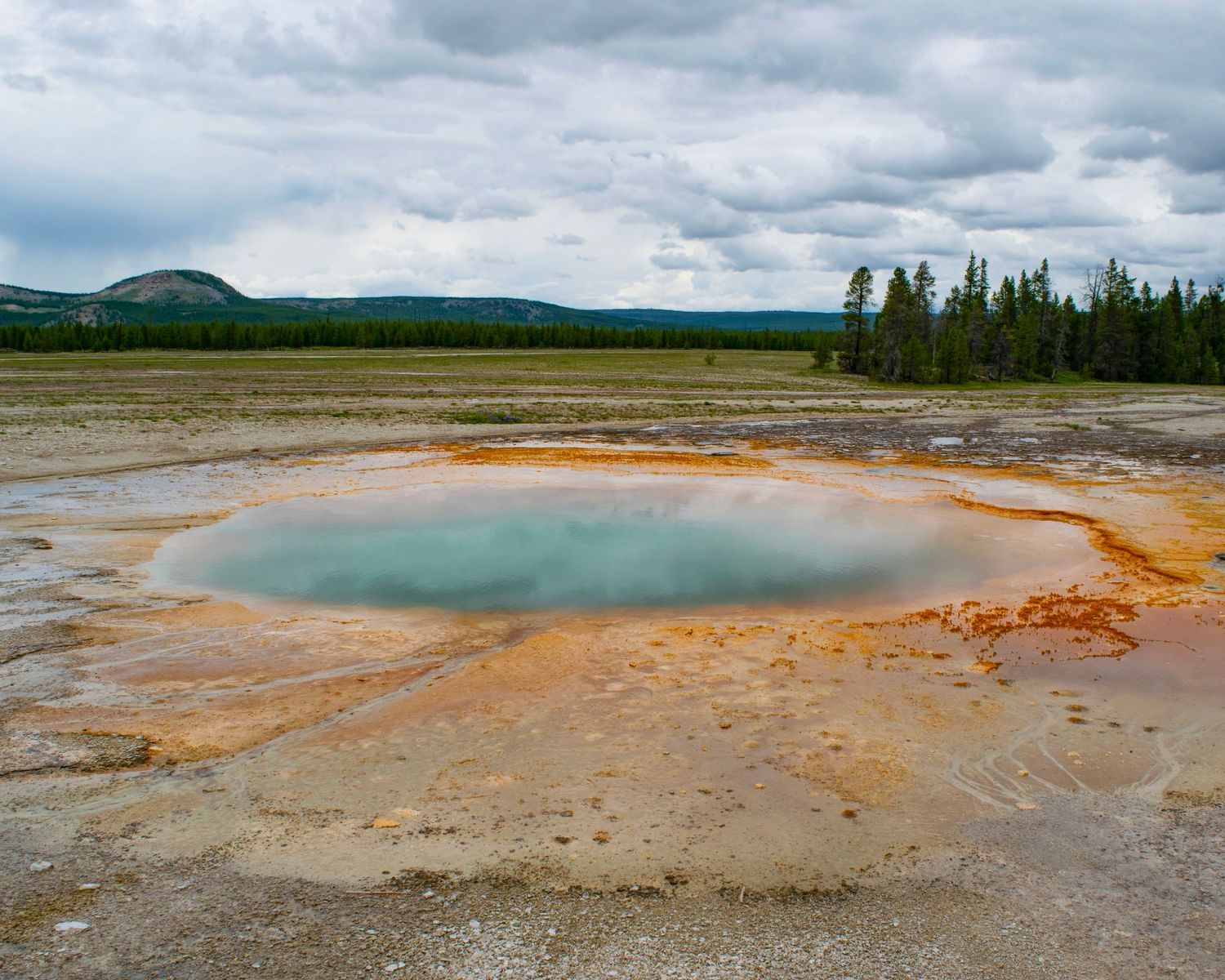 Colorful hot spring pool with orange mineral deposits at Yellowstone National Park under cloudy skies.