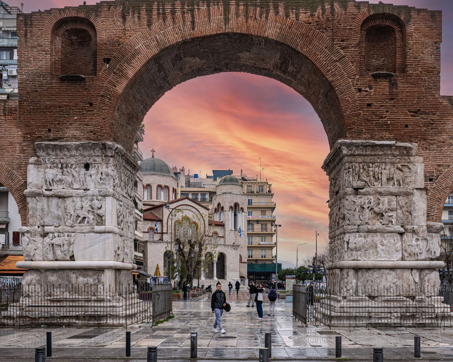 Ancient Roman arch framing a Byzantine church at sunset in Thessaloniki, Greece, with warm pink skies.