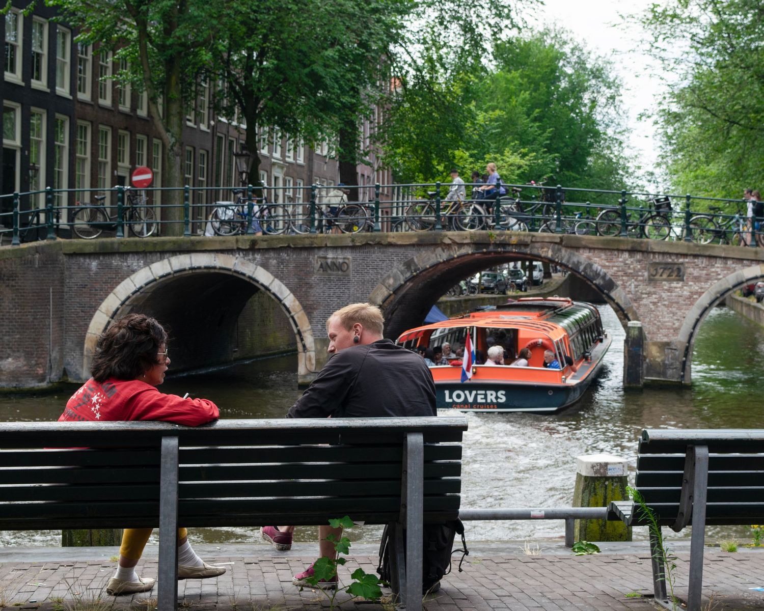 Couple sitting on bench by Amsterdam canal watching a boat named Lovers pass under a stone bridge.