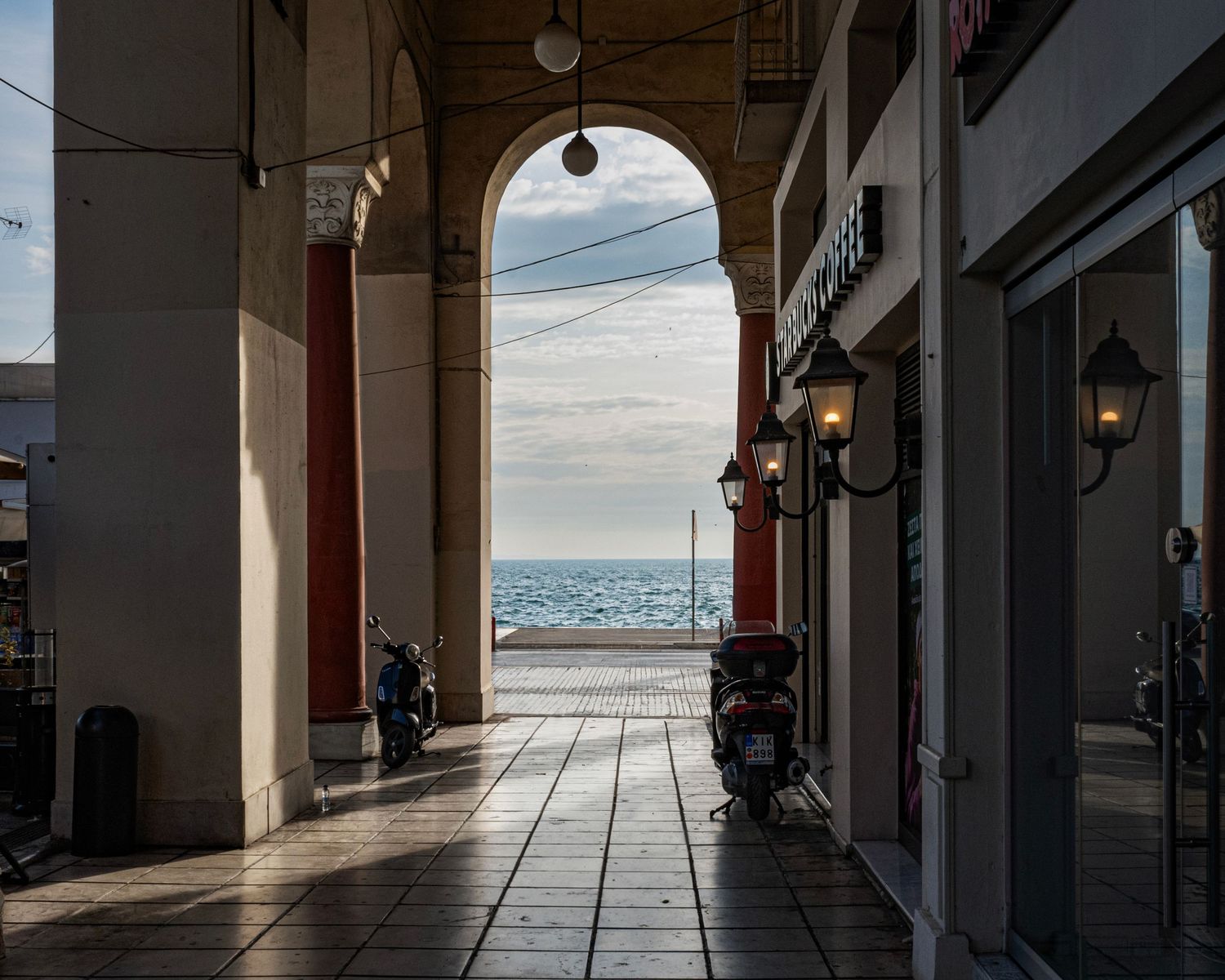 A covered archway passage with stone columns frames a serene ocean view, with vintage lanterns and parked scooters nearby.