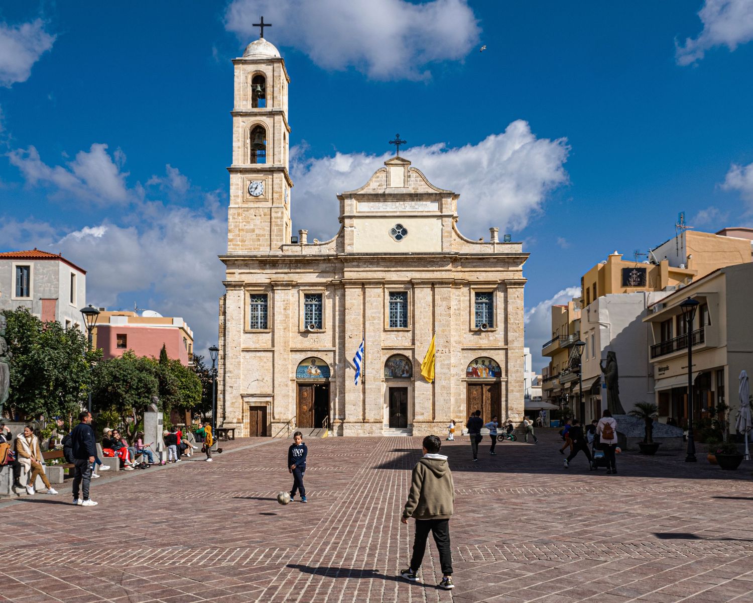 Historic cathedral with bell tower on a sunny plaza in Chania, Crete, with visitors and Greek flag.