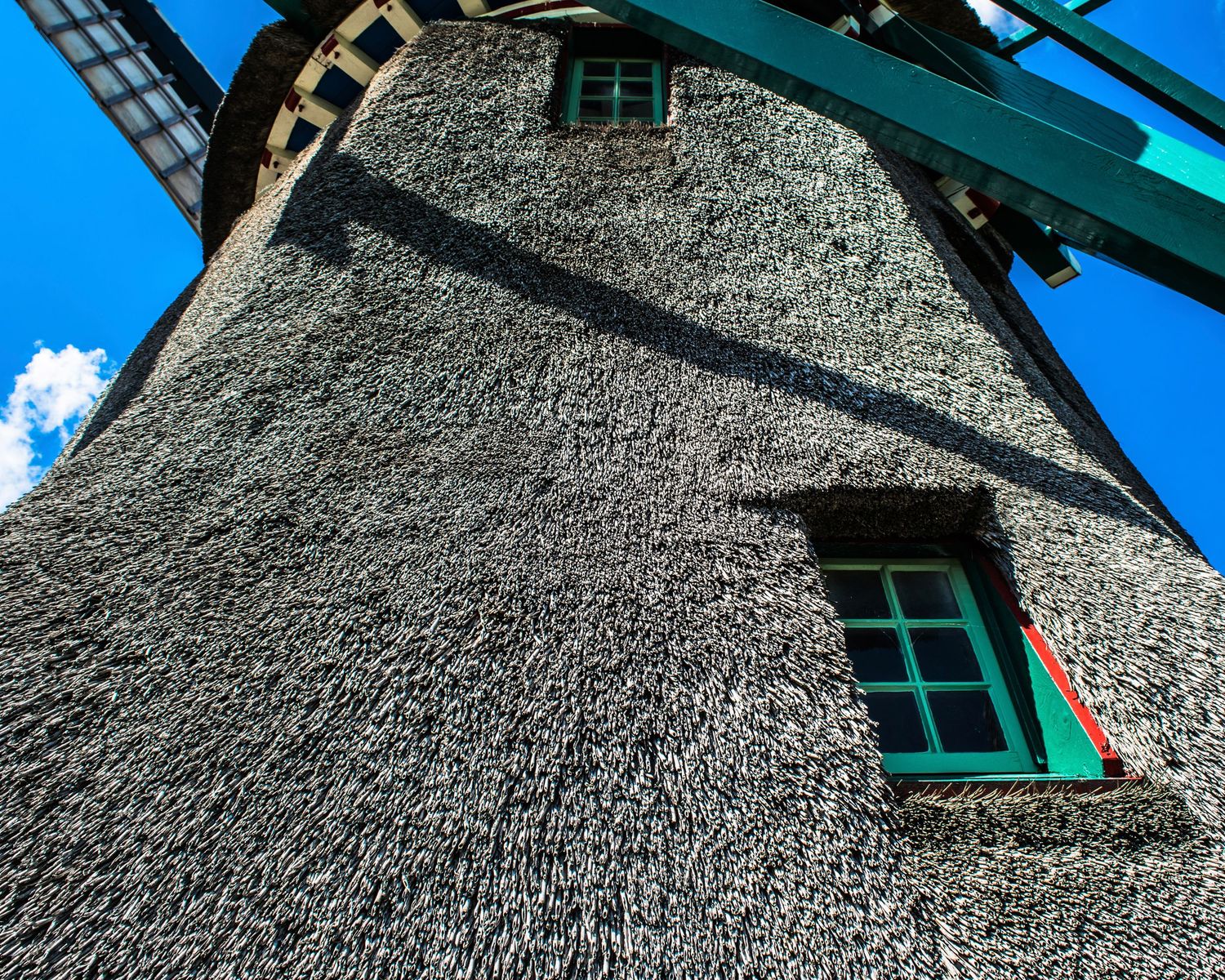 Historic Dutch windmill with thatched exterior, green-framed windows, and wooden sails against a bright blue sky.