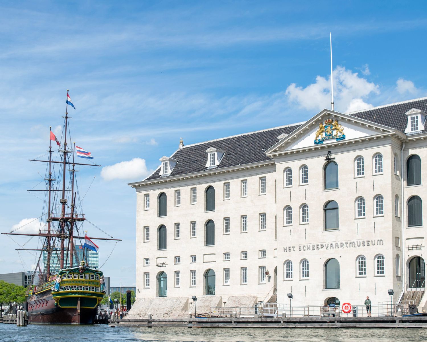 Historic Dutch maritime museum with tall ship replica docked alongside classical white building in Amsterdam waterfront.