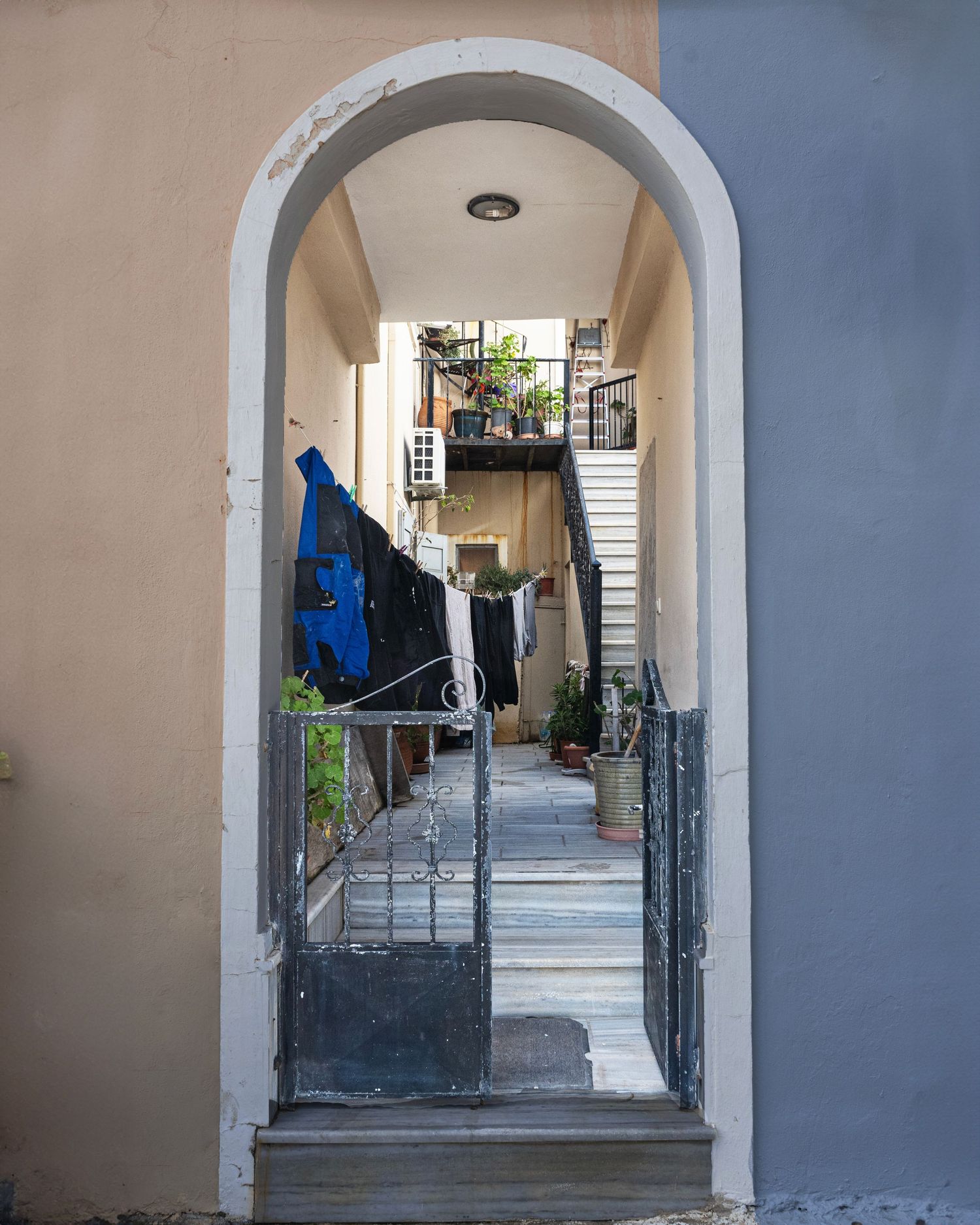 Arched doorway with iron gate leads to a narrow alleyway with steps, plants, and laundry in an urban building.