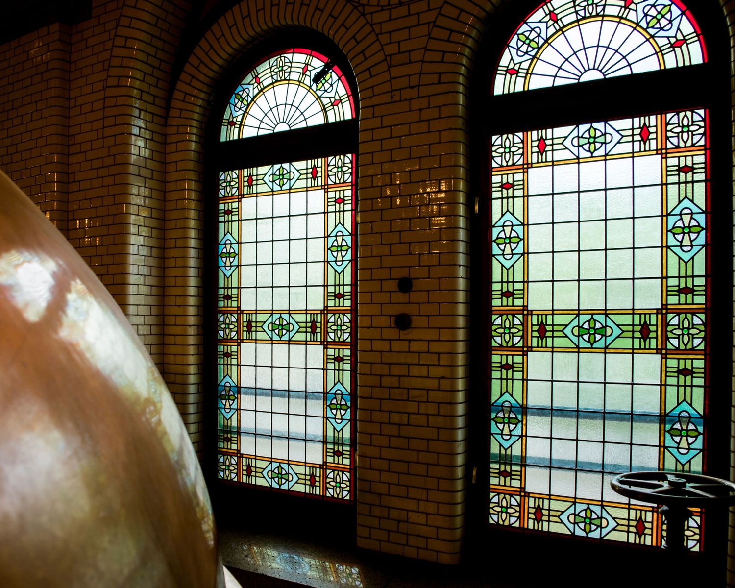 Ornate stained glass windows with geometric patterns in green and gold, set in brick arches beside a copper vessel.