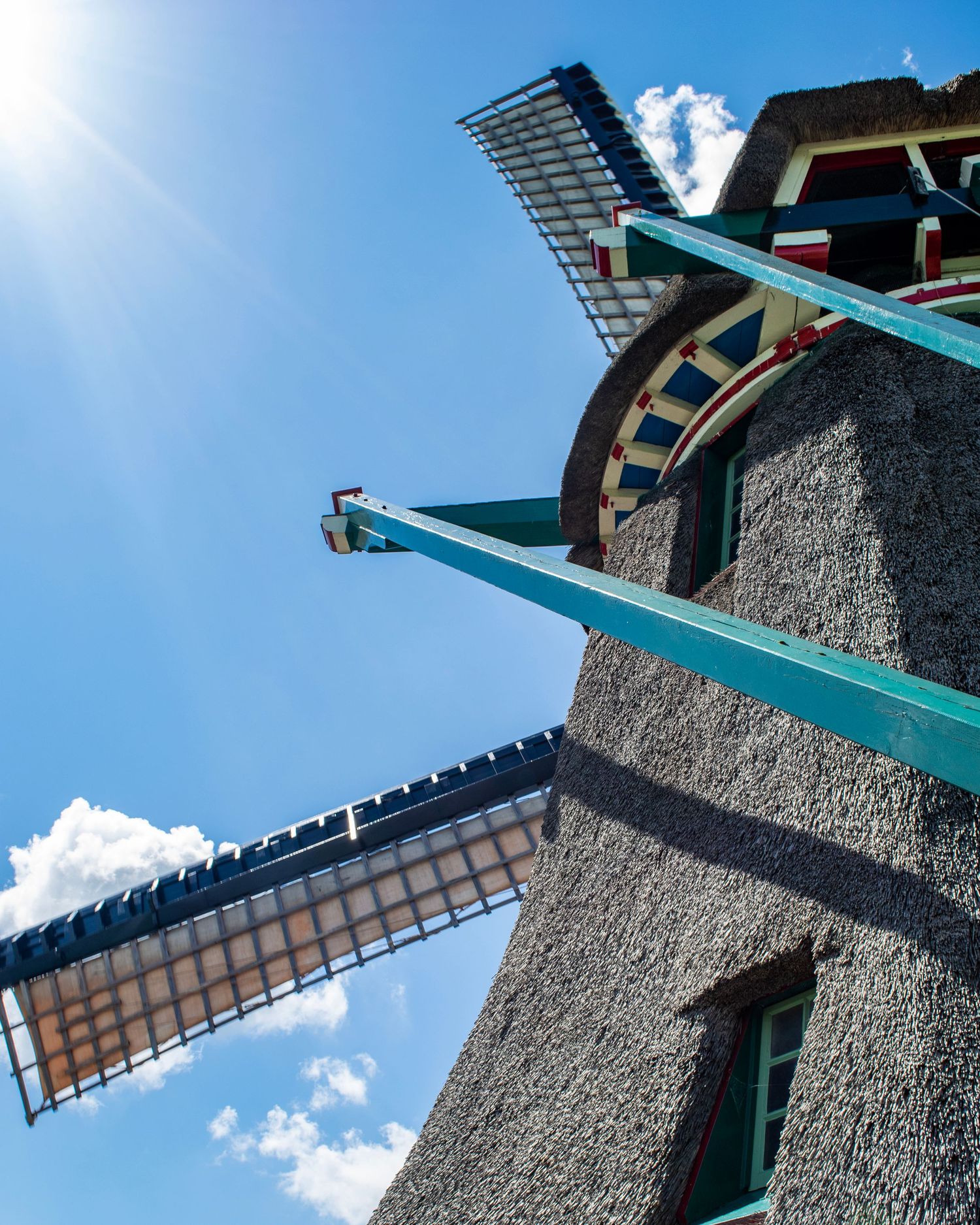 Low-angle view of a traditional Dutch windmill with teal beams and stone exterior against a bright blue sky.