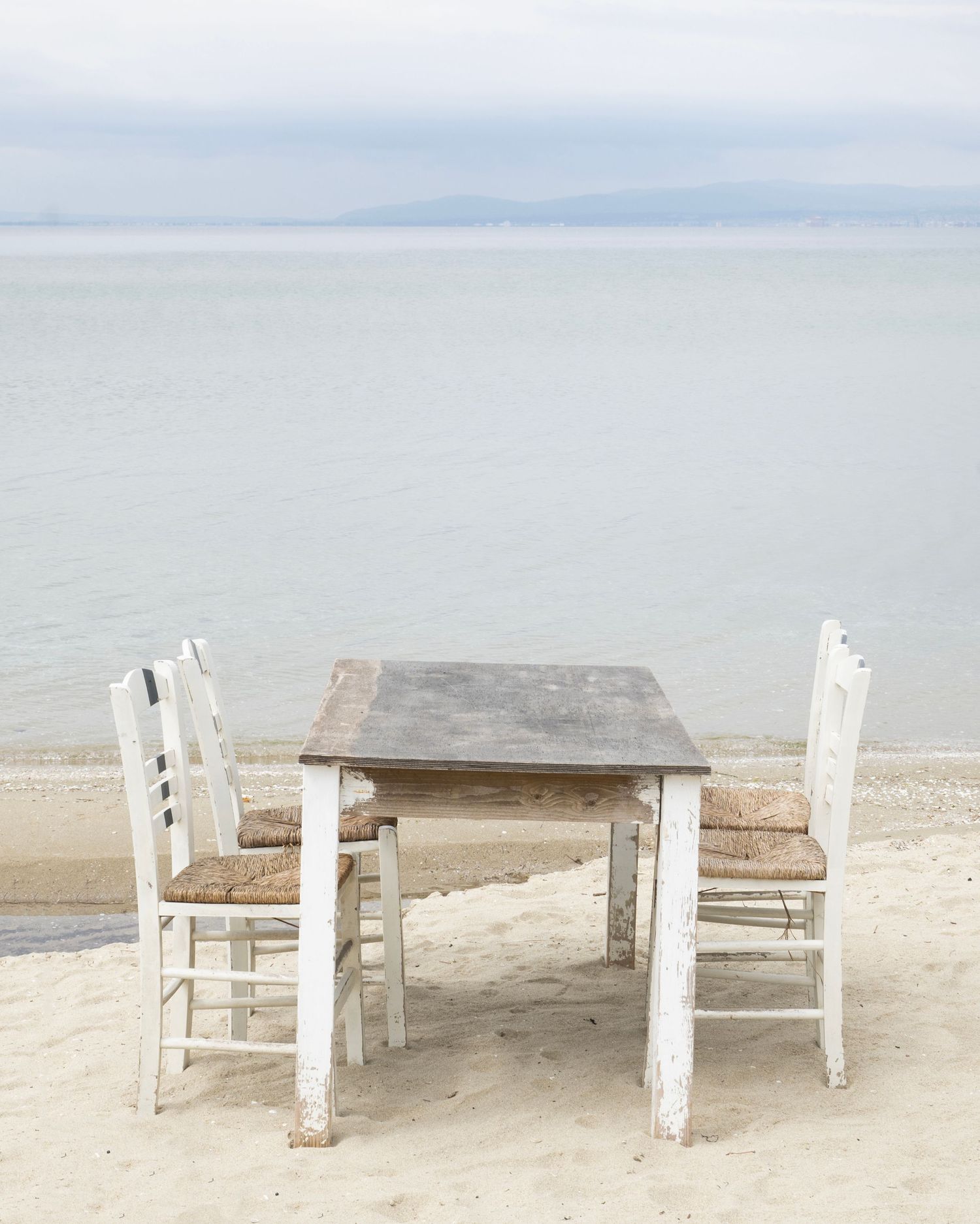 Rustic wooden table with two white chairs on a sandy beach overlooking a calm sea and distant mountains.