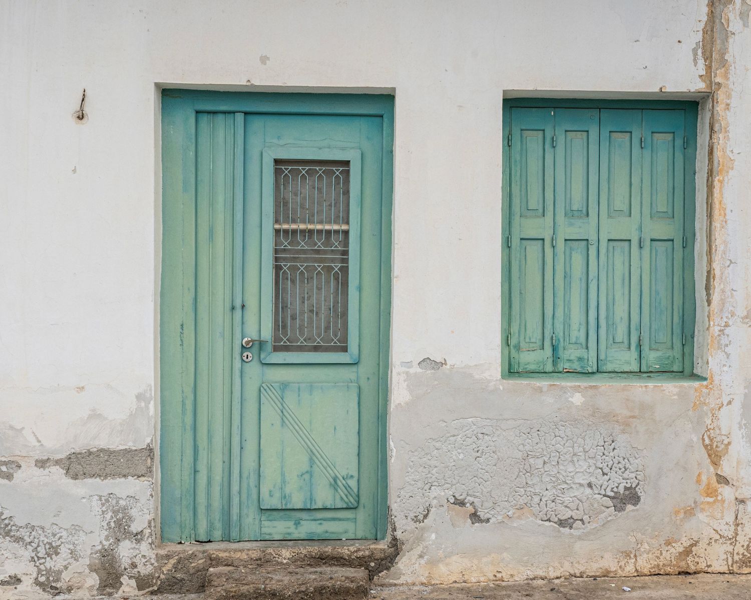 Weathered white building facade with turquoise green wooden door and shuttered window, rustic Mediterranean architecture.